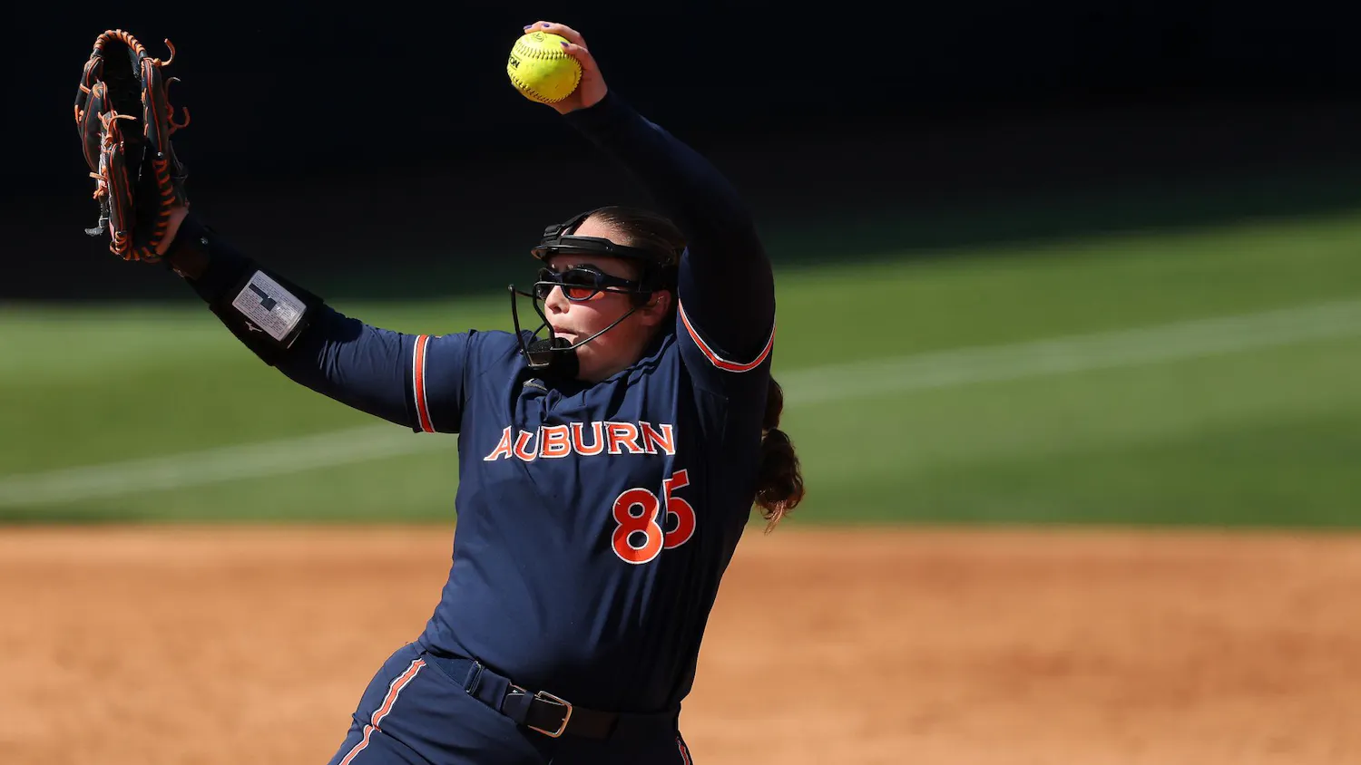 A young female softball player in a dark blue uniform winds up to pitch, holding a bright yellow softball in her right hand.