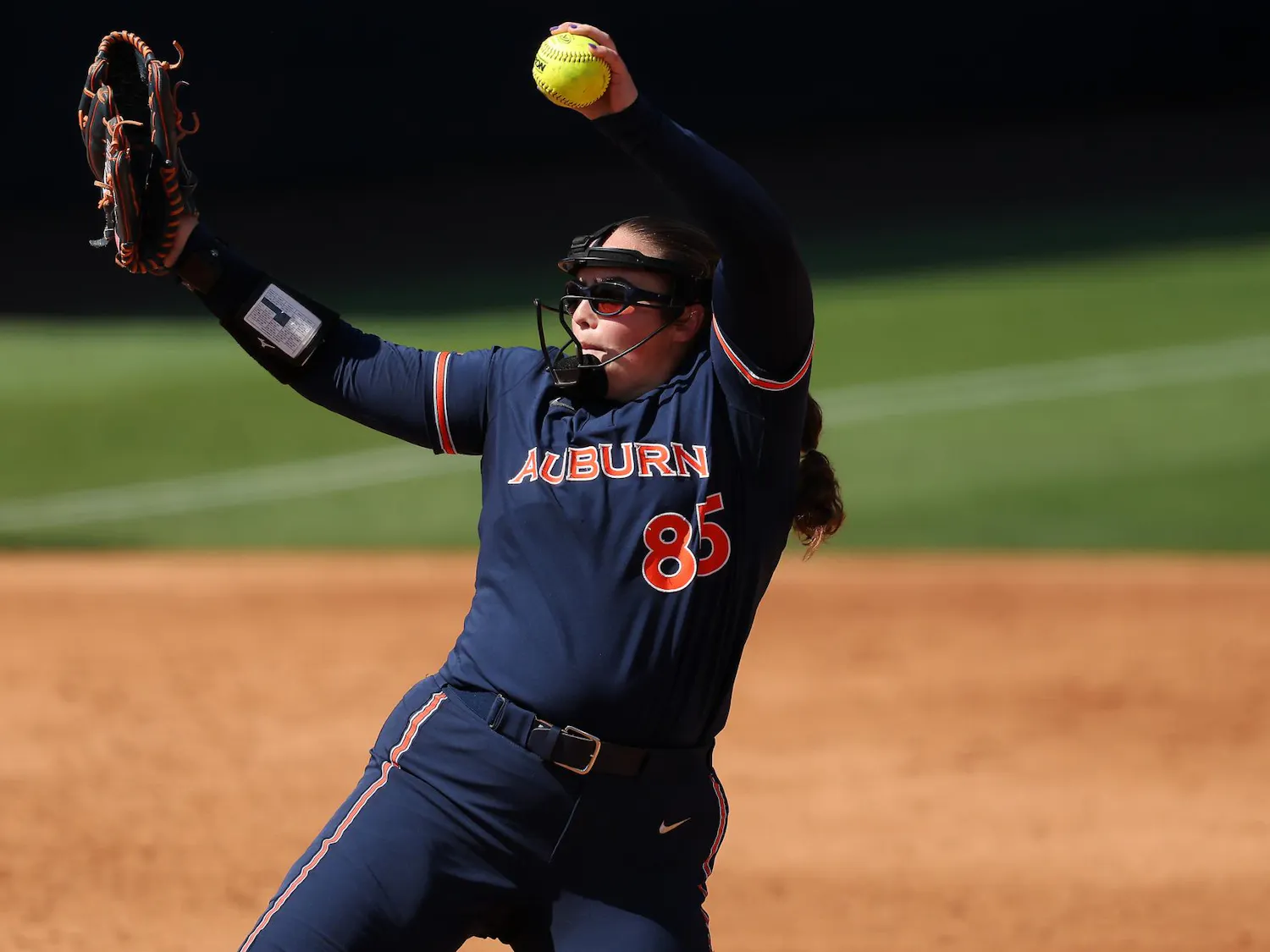 A young female softball player in a dark blue uniform winds up to pitch, holding a bright yellow softball in her right hand.
