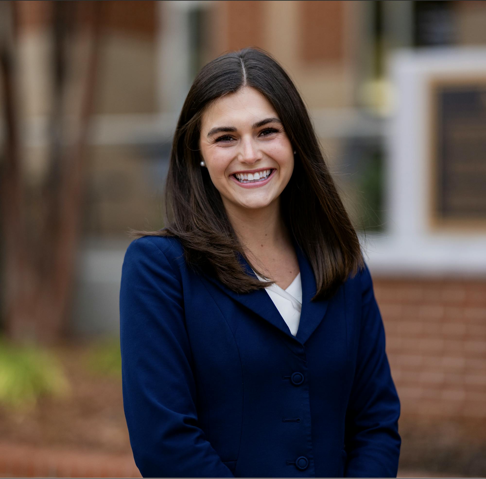 A girl with dark brown hair with a blue blazer is smiling, with a blurred background of a brick building and a tree. 