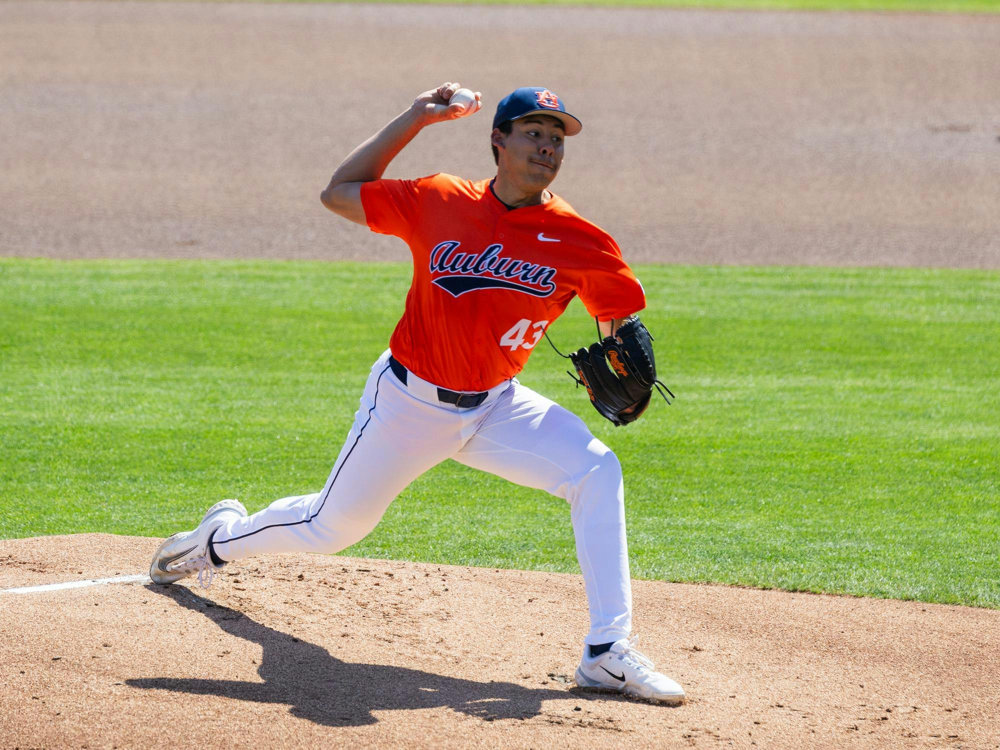 A male baseball pitcher wearing an orange jersey and white pants is mid-delivery on a grassy field.
