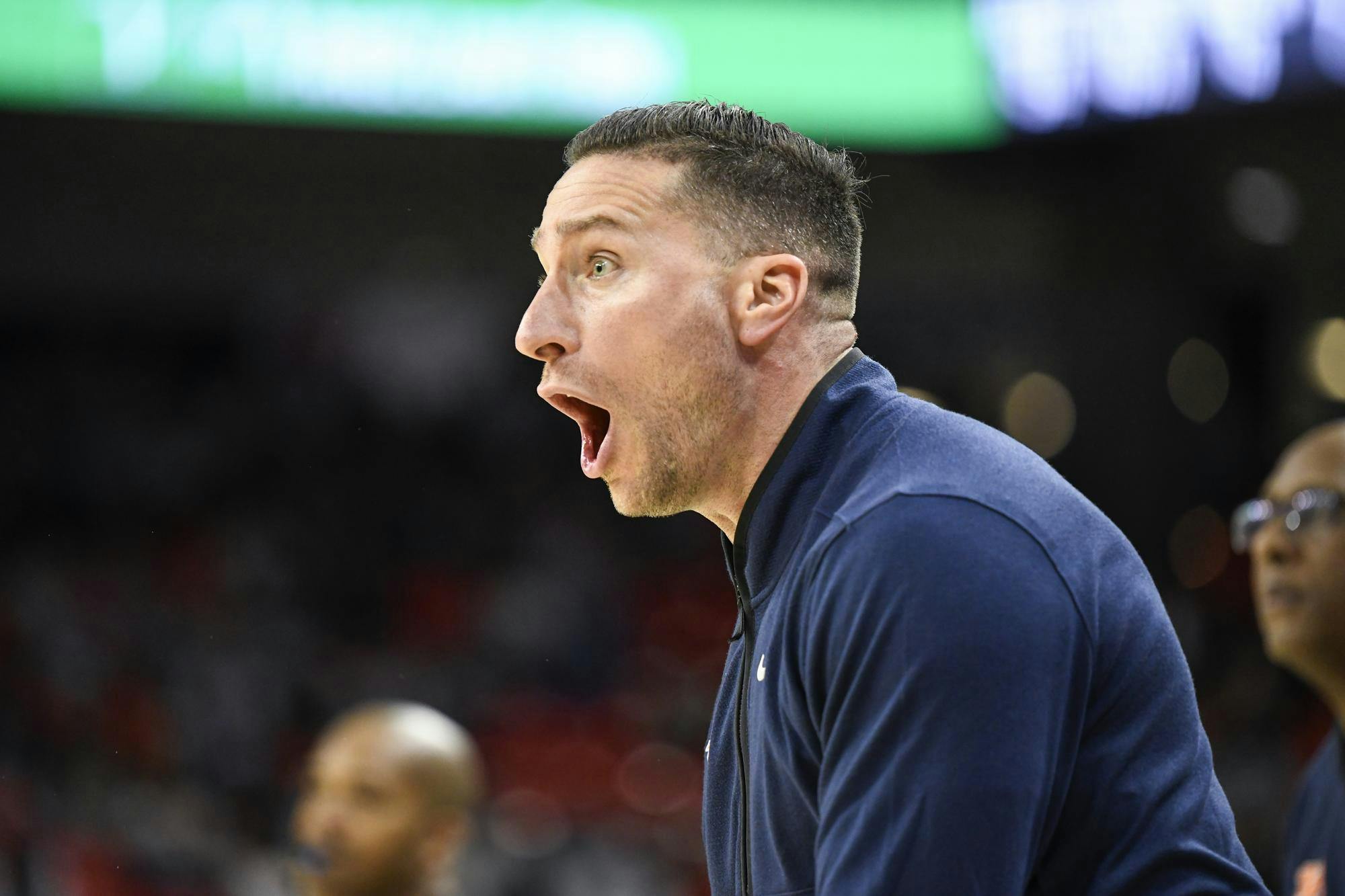 A man with short, slicked-back hair is animatedly shouting, his mouth wide open, while wearing a dark blue jacket on a blurred background.