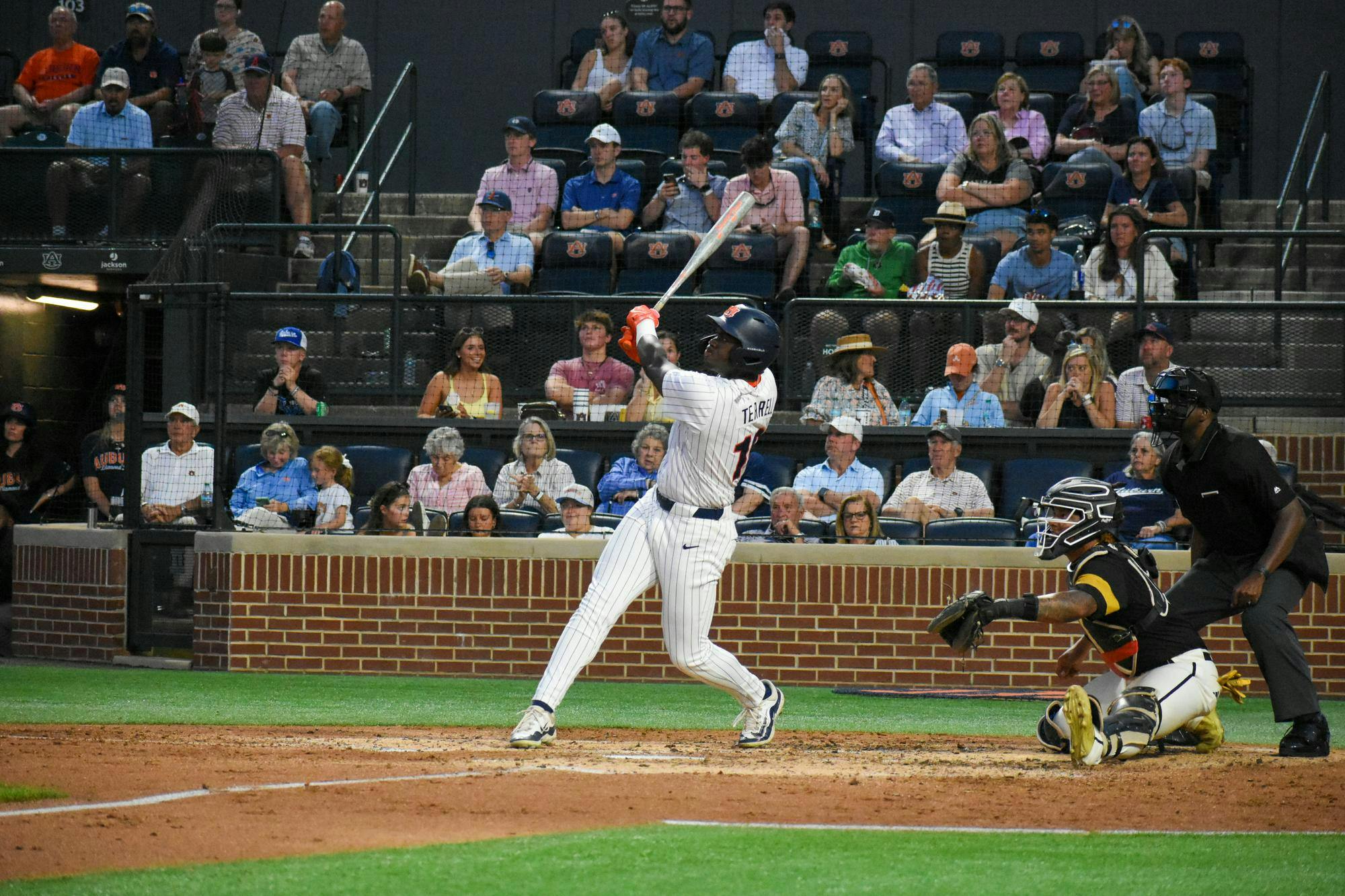 A baseball player swings a bat while a catcher crouches behind home plate, with spectators watching in the background.