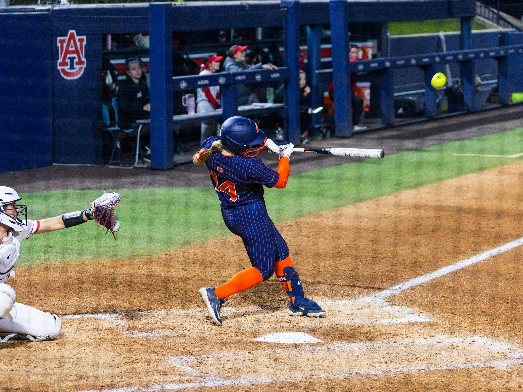 A player in a navy and orange uniform swings a bat at a softball as a catcher in white crouches behind home plate.