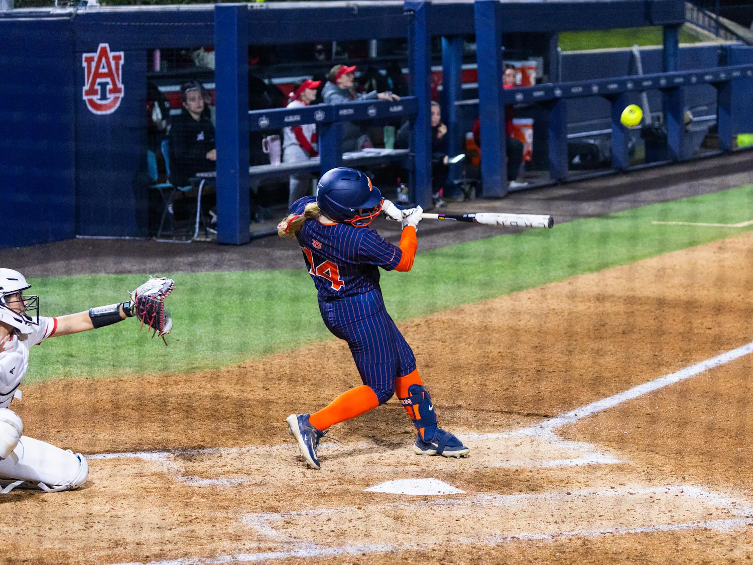 A player in a navy and orange uniform swings a bat at a softball as a catcher in white crouches behind home plate.