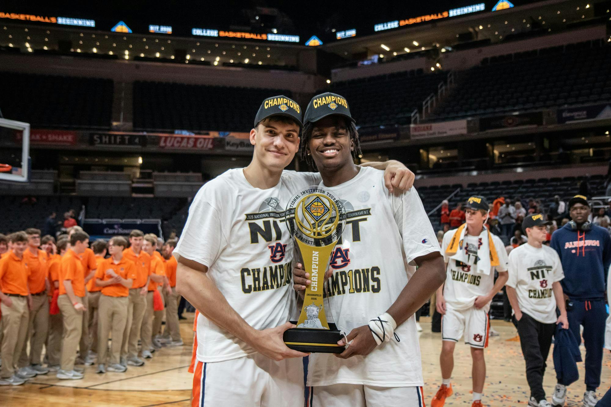 Two young men, wearing white shirts and black "Champions" hats, hold a trophy in a basketball arena with fans in the background.