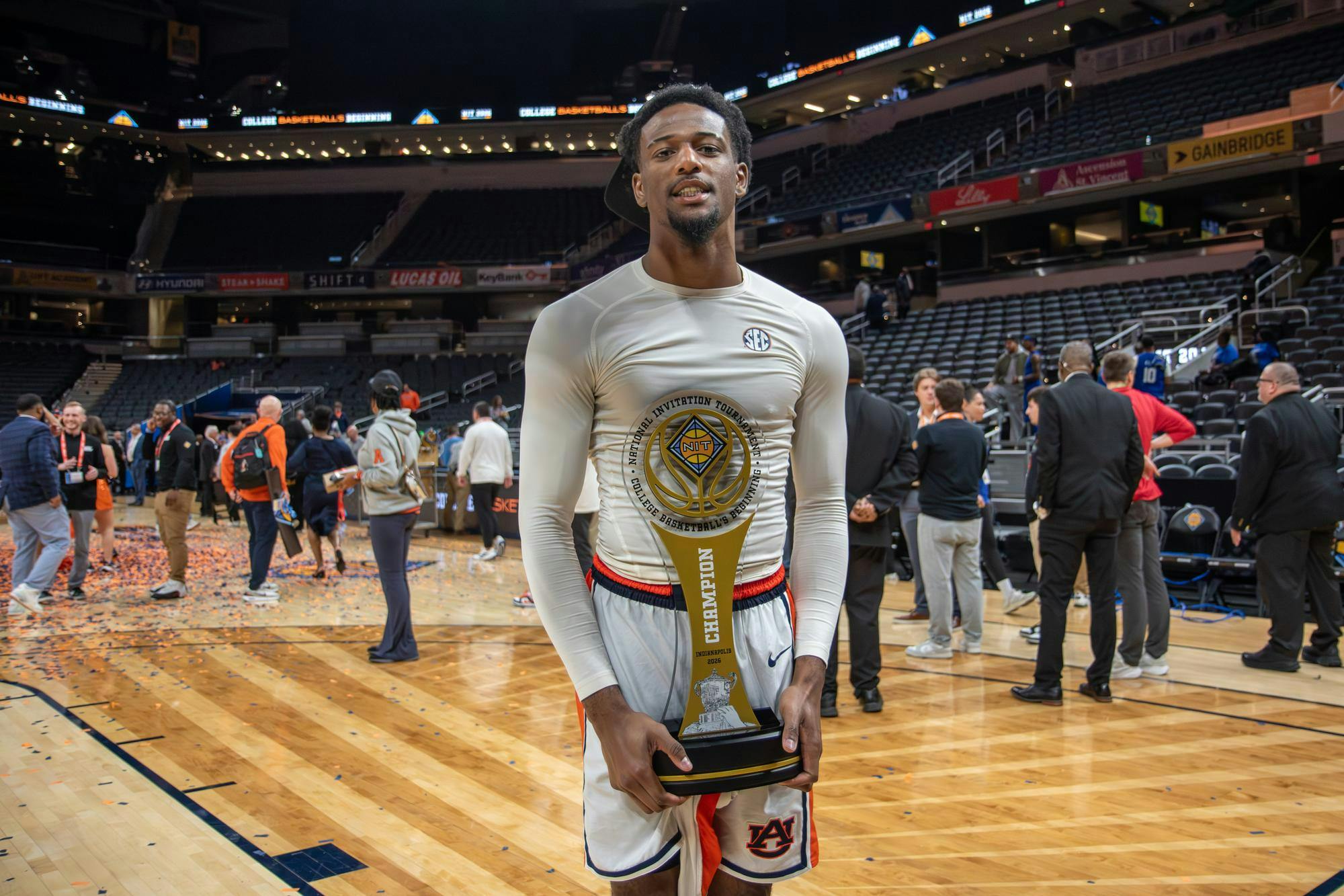 A basketball player in a white long-sleeve shirt stands on a court, holding a large trophy amidst a celebratory crowd.