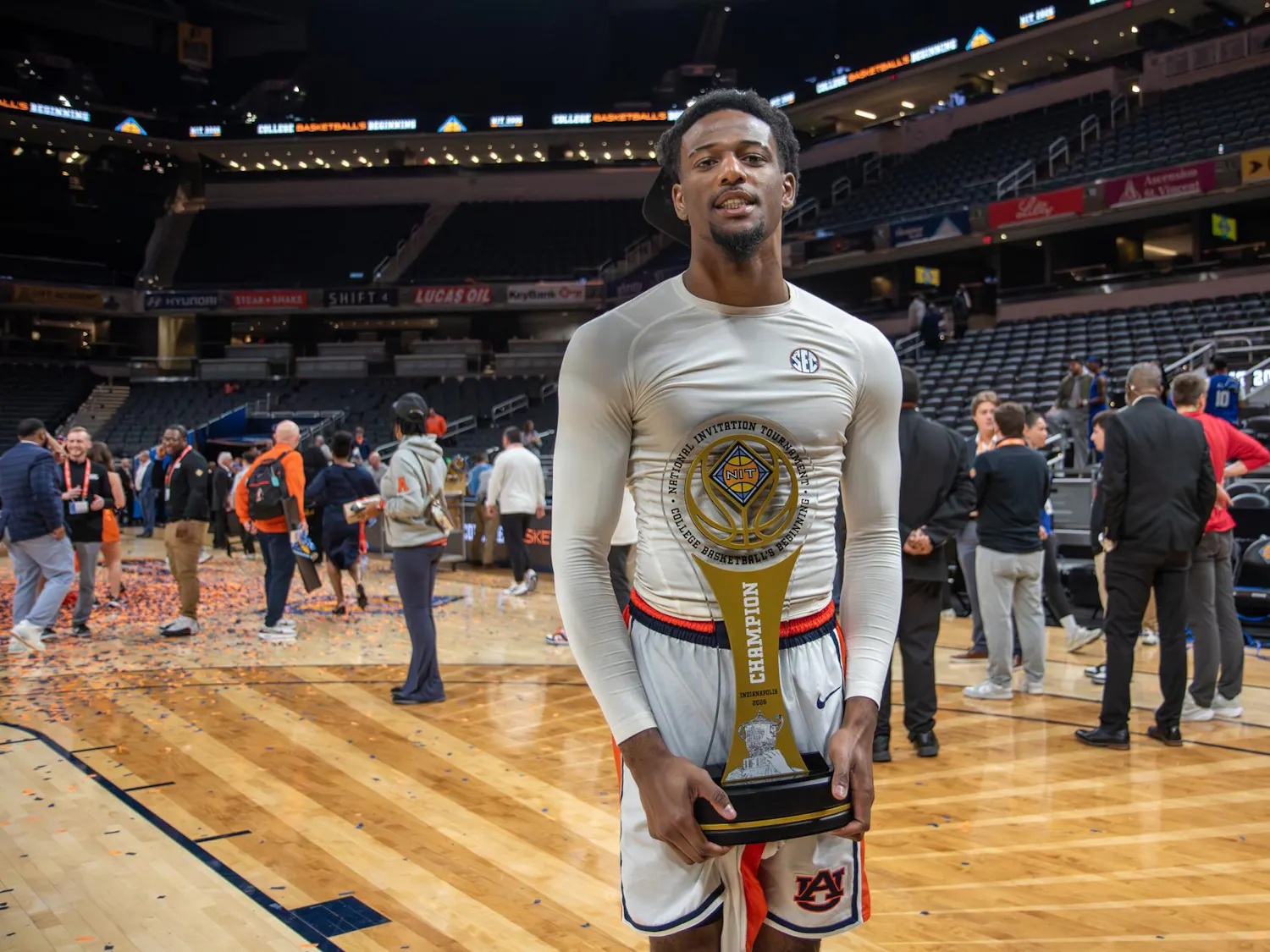 A basketball player in a white long-sleeve shirt stands on a court, holding a large trophy amidst a celebratory crowd.