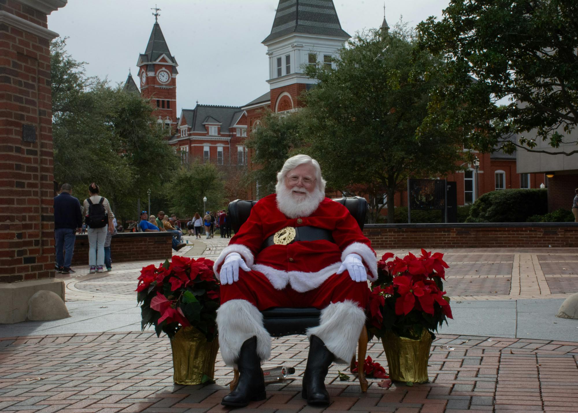 Santa Claus sits on a chair surrounded by poinsettias, with a campus backdrop and people walking by.