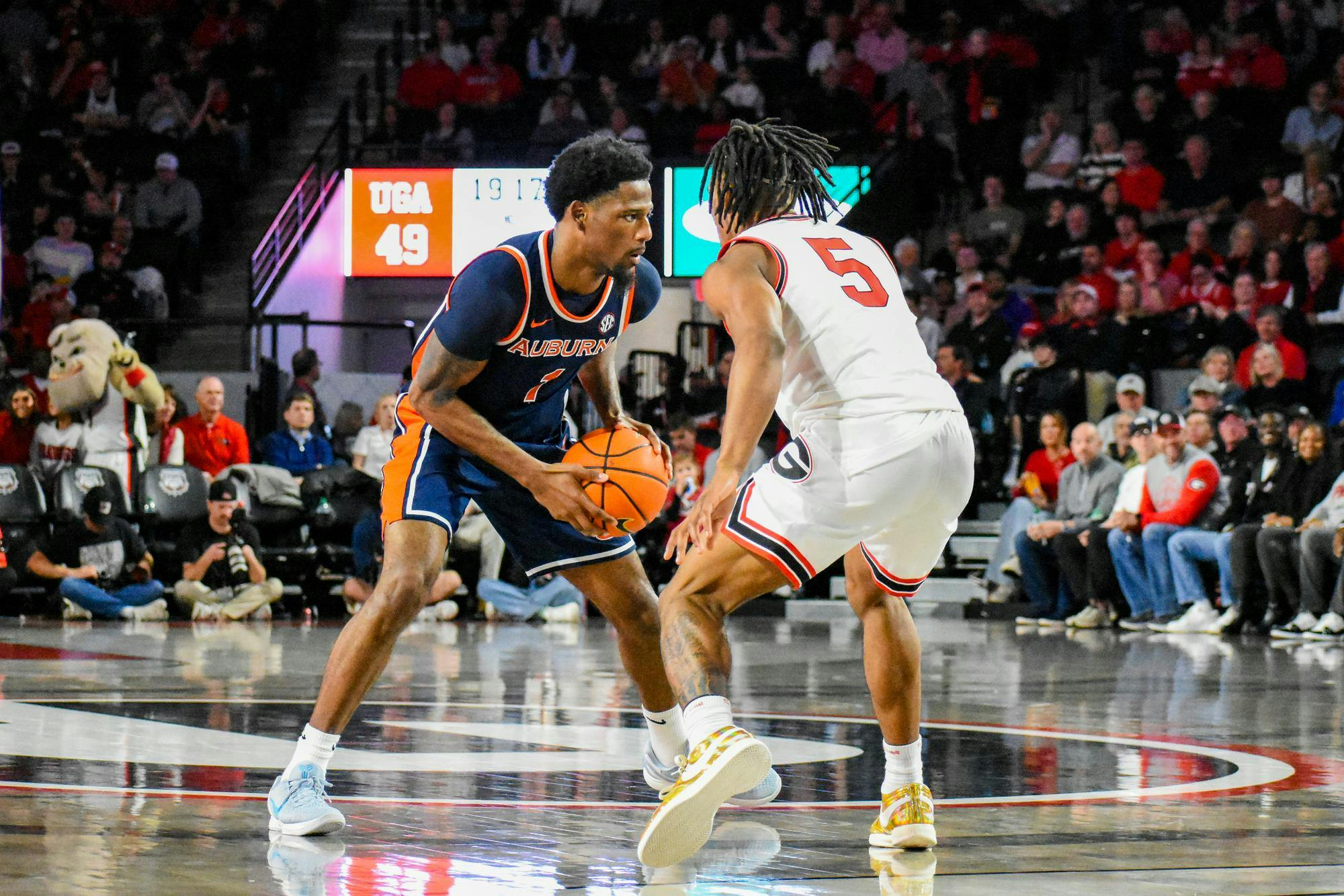 Two basketball players face each other on the court, one dribbling a ball while a cheering crowd fills the background.