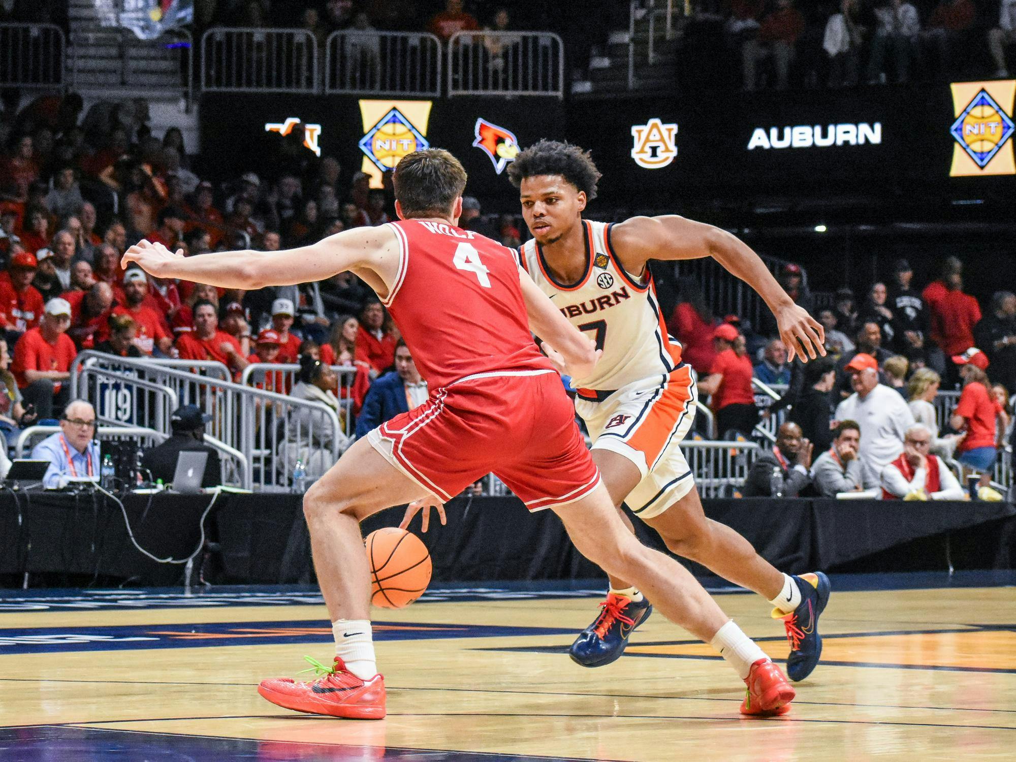 Two basketball players are engaged in a game, one dribbling a ball while the other defends closely; a crowd is visible in the background.