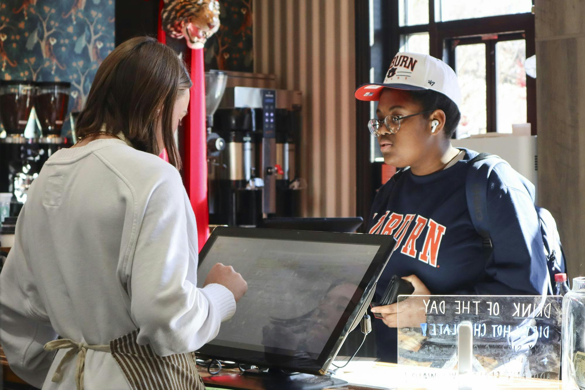 A customer wearing a baseball cap interacts with a barista at a café, both standing near a touchscreen register.