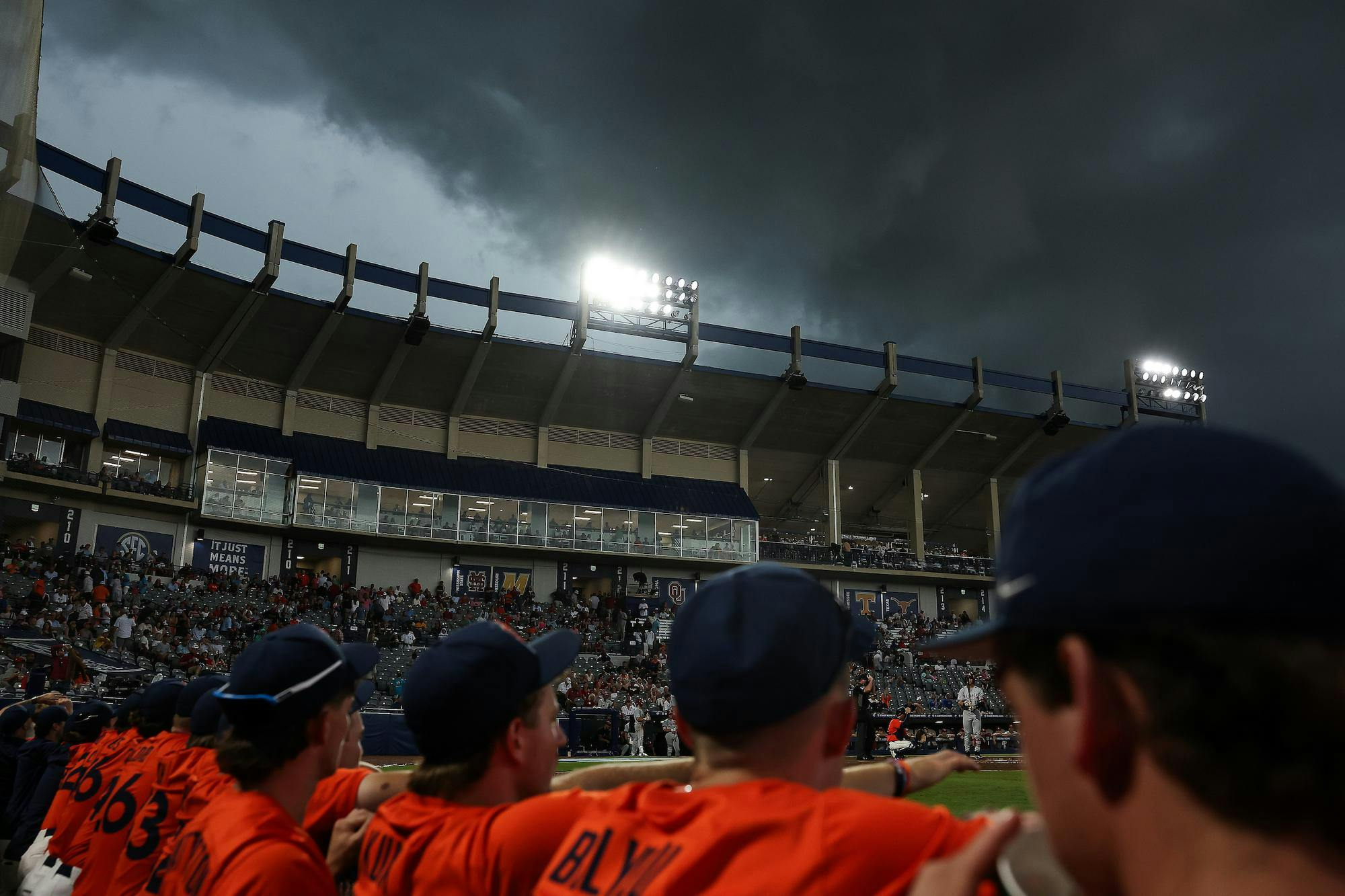 A group of players in orange uniforms stands with their backs to the viewer, while dark storm clouds loom over a stadium filled with spectators.