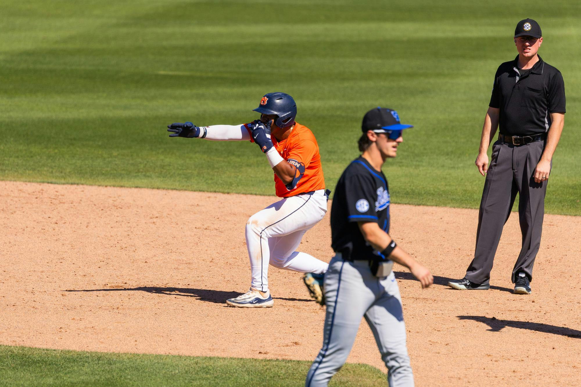 A baseball player in an orange jersey appears to celebrate while another player in black walks by, with an umpire observing nearby.