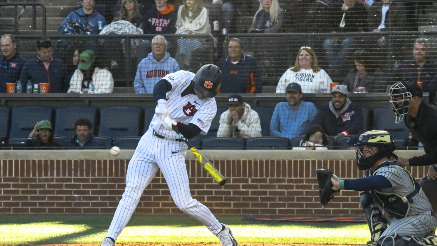 A baseball player in a pinstriped uniform swings a bat at a pitch, with spectators watching in the background.