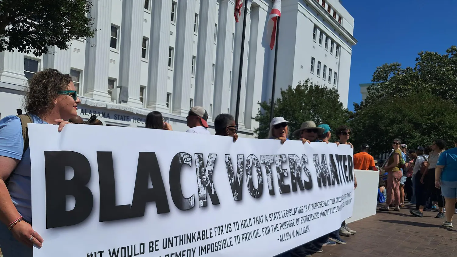 Protesters stand outside the Alabama State House and hold a sign reading “Black Voters Matter” with a quote from the Allen v. Milligan case under it.