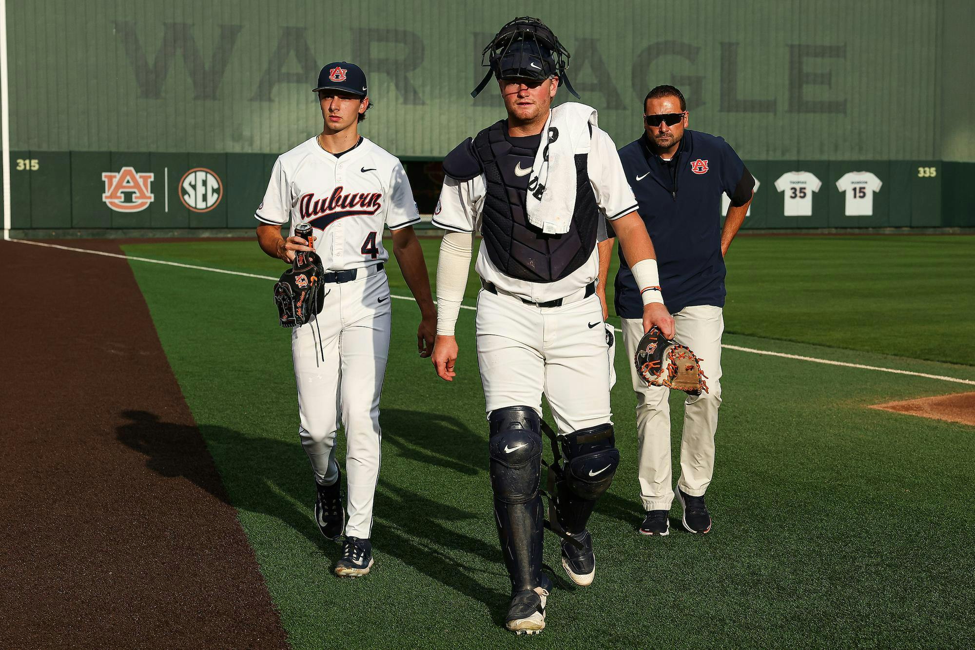 Two baseball players in white uniforms walk on a field, accompanied by a coach, with a large wall displaying “WAR EAGLE” behind them.