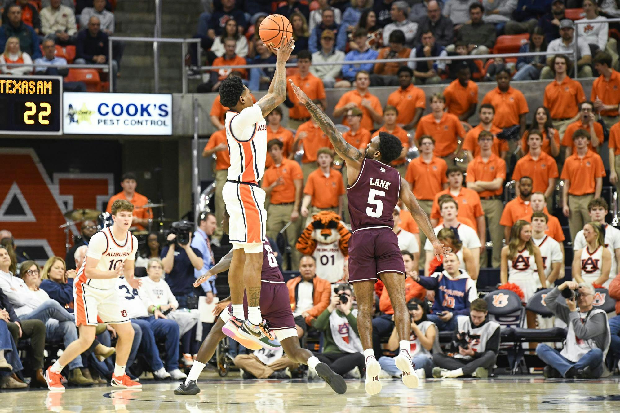 A player in an orange and white uniform jumps to shoot a basketball while being defended by an opponent in maroon.