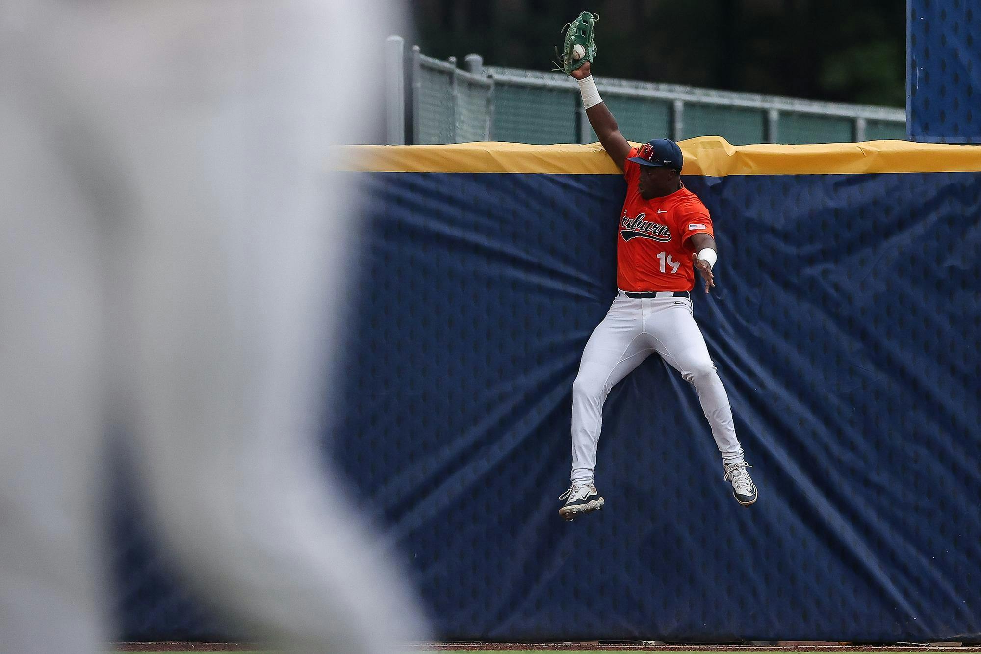 A baseball player in an orange jersey leaps against a blue wall, reaching for a ball with one hand.