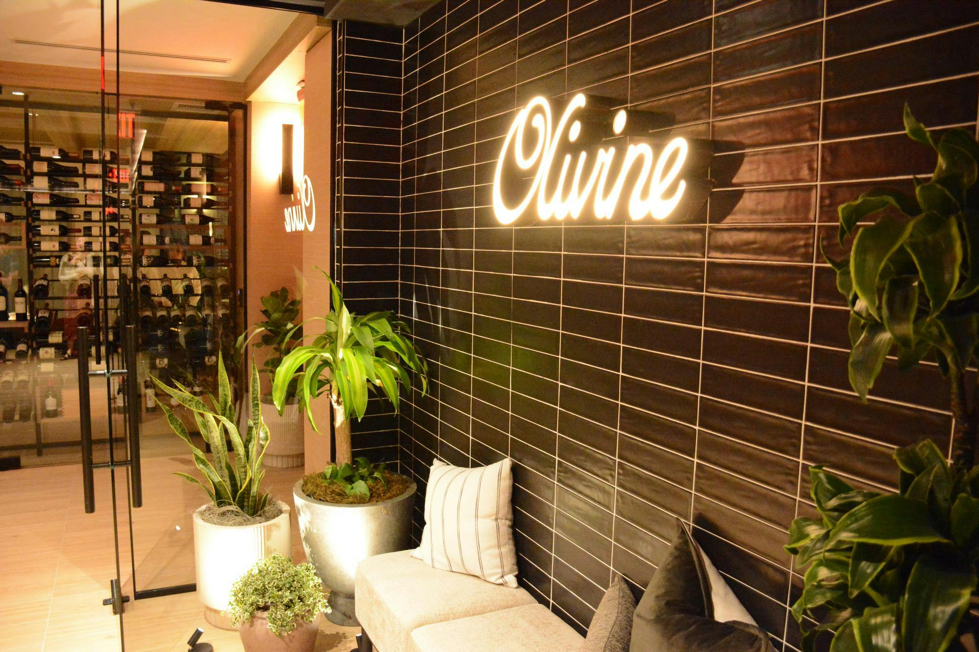 A stylish interior features a dark tiled wall, illuminated sign reading "Olivine," lush green plants, and plush seating.
