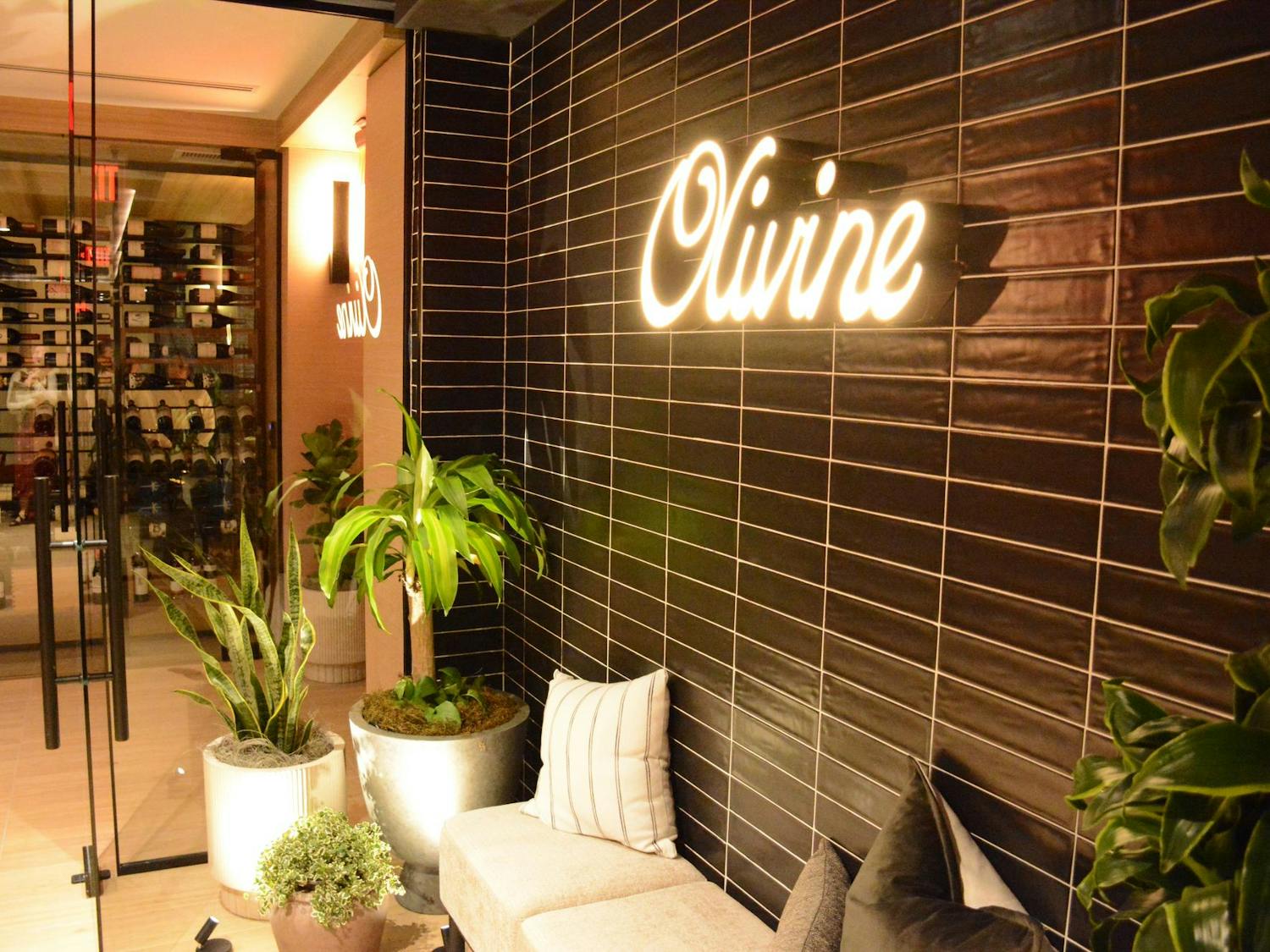 A stylish interior features a dark tiled wall, illuminated sign reading "Olivine," lush green plants, and plush seating.