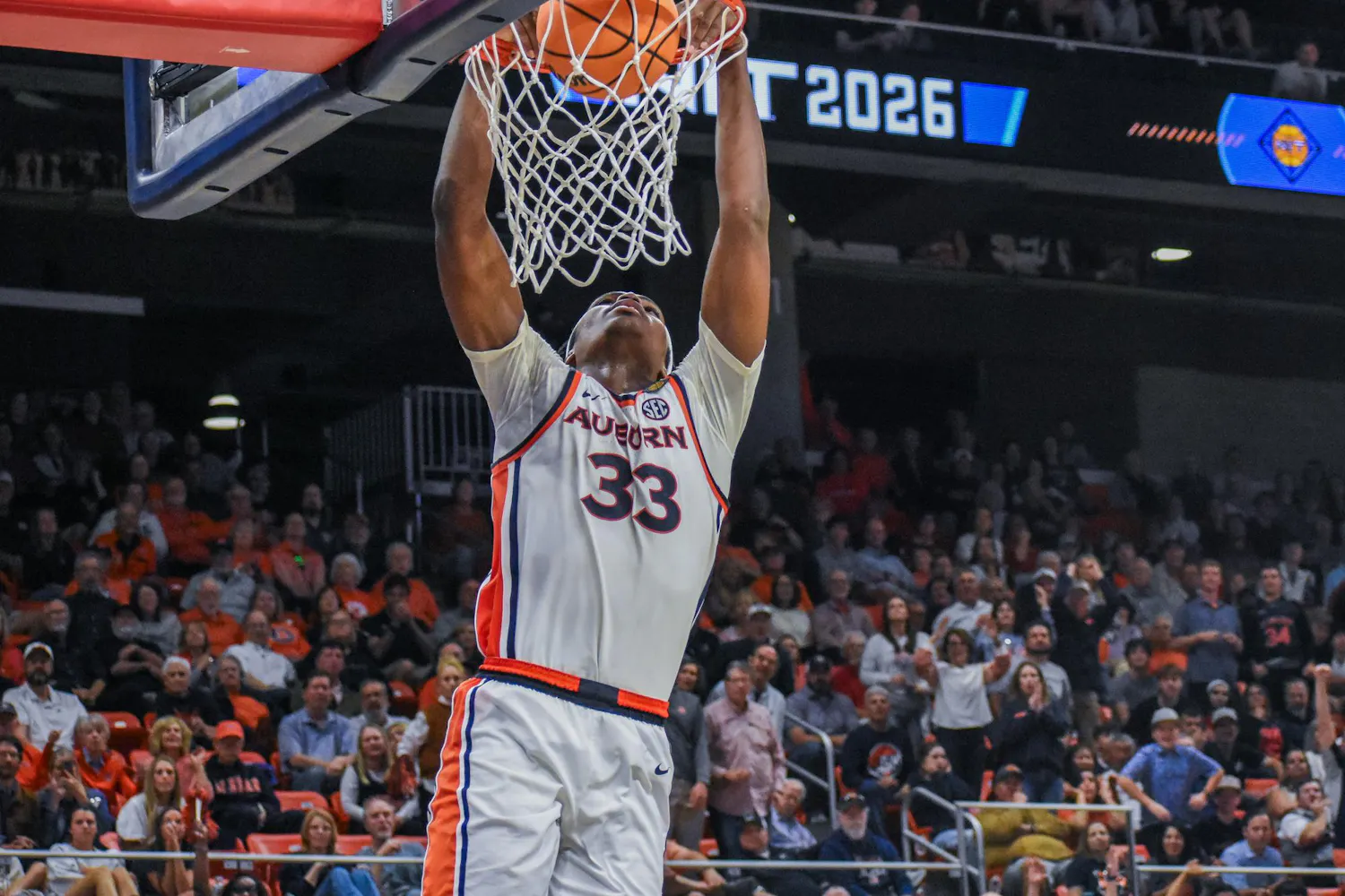 A basketball player in a white uniform with orange and blue accents is performing a dunk into the hoop.