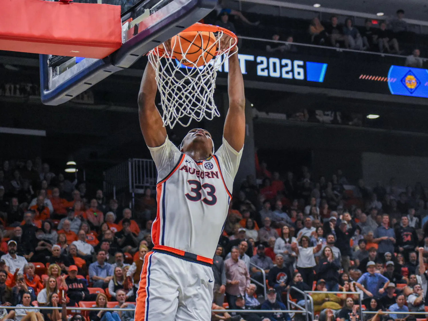 A basketball player in a white uniform with orange and blue accents is performing a dunk into the hoop.