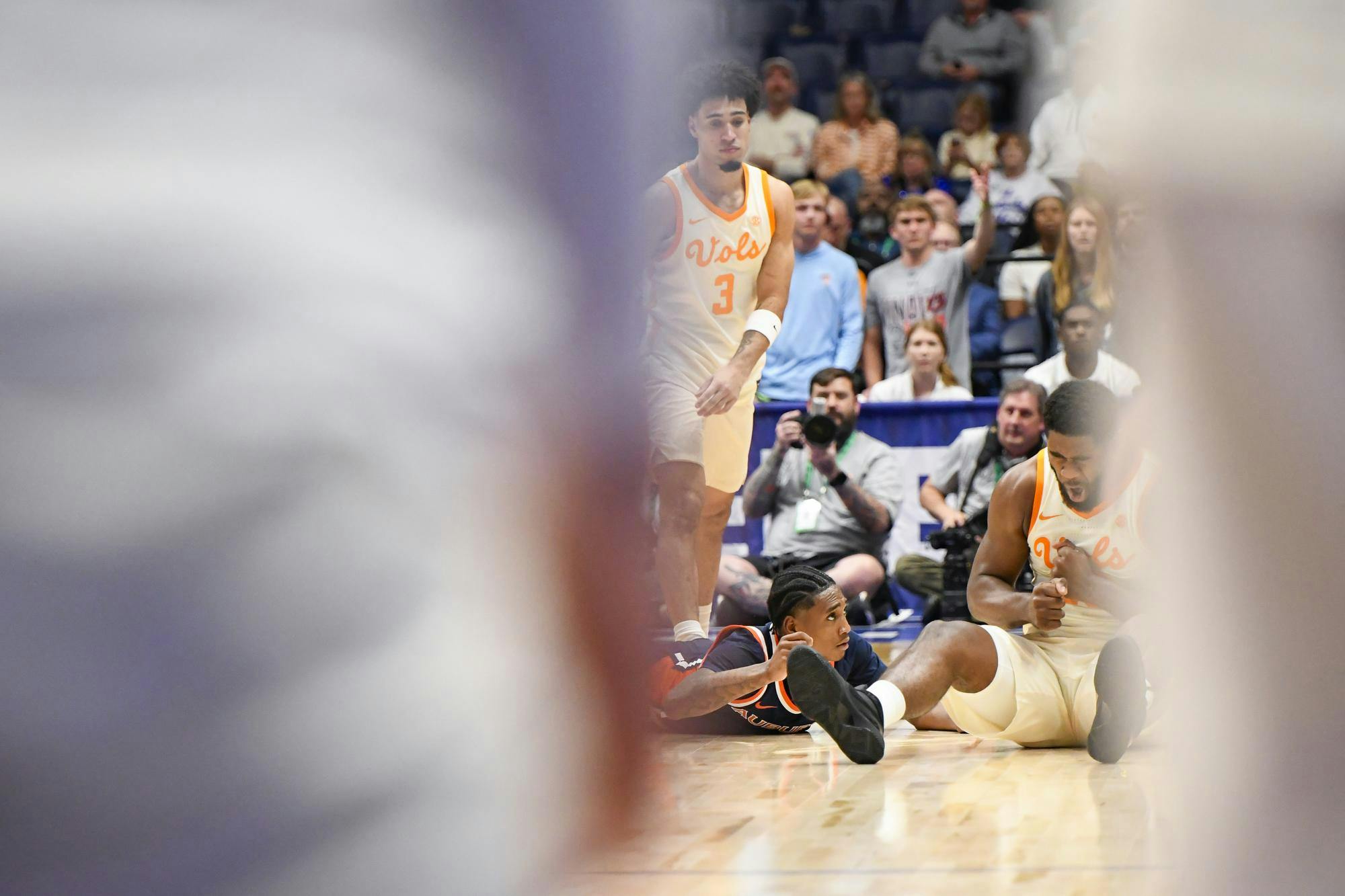 Two basketball players are on the court, one lying down and another standing, with a crowd watching in the background.