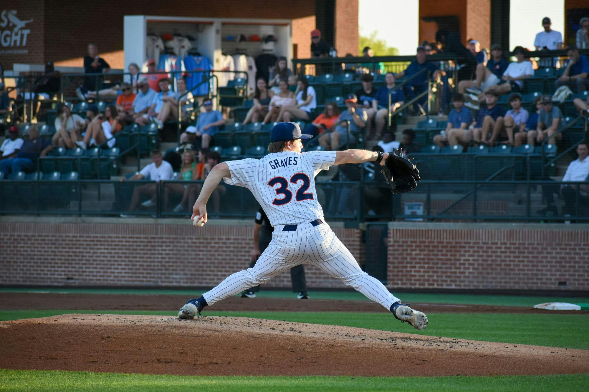 A baseball pitcher in a striped uniform is in mid-throw on the pitcher's mound, with spectators in the background.