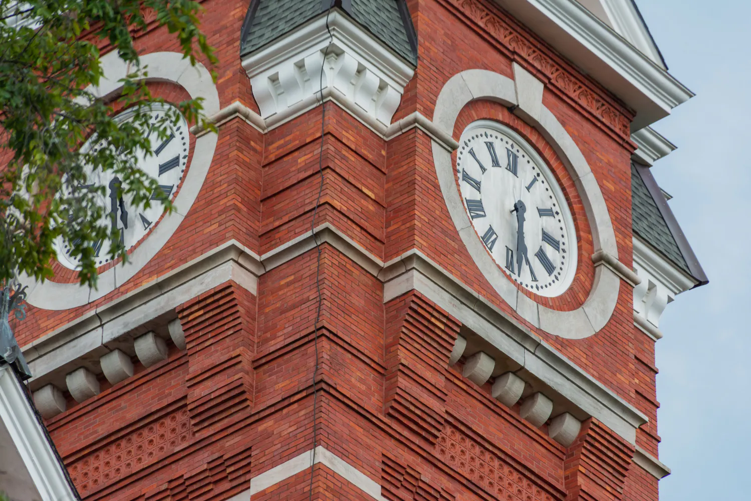 A red brick clock tower features two large round clocks with Roman numerals and a pointed roof.