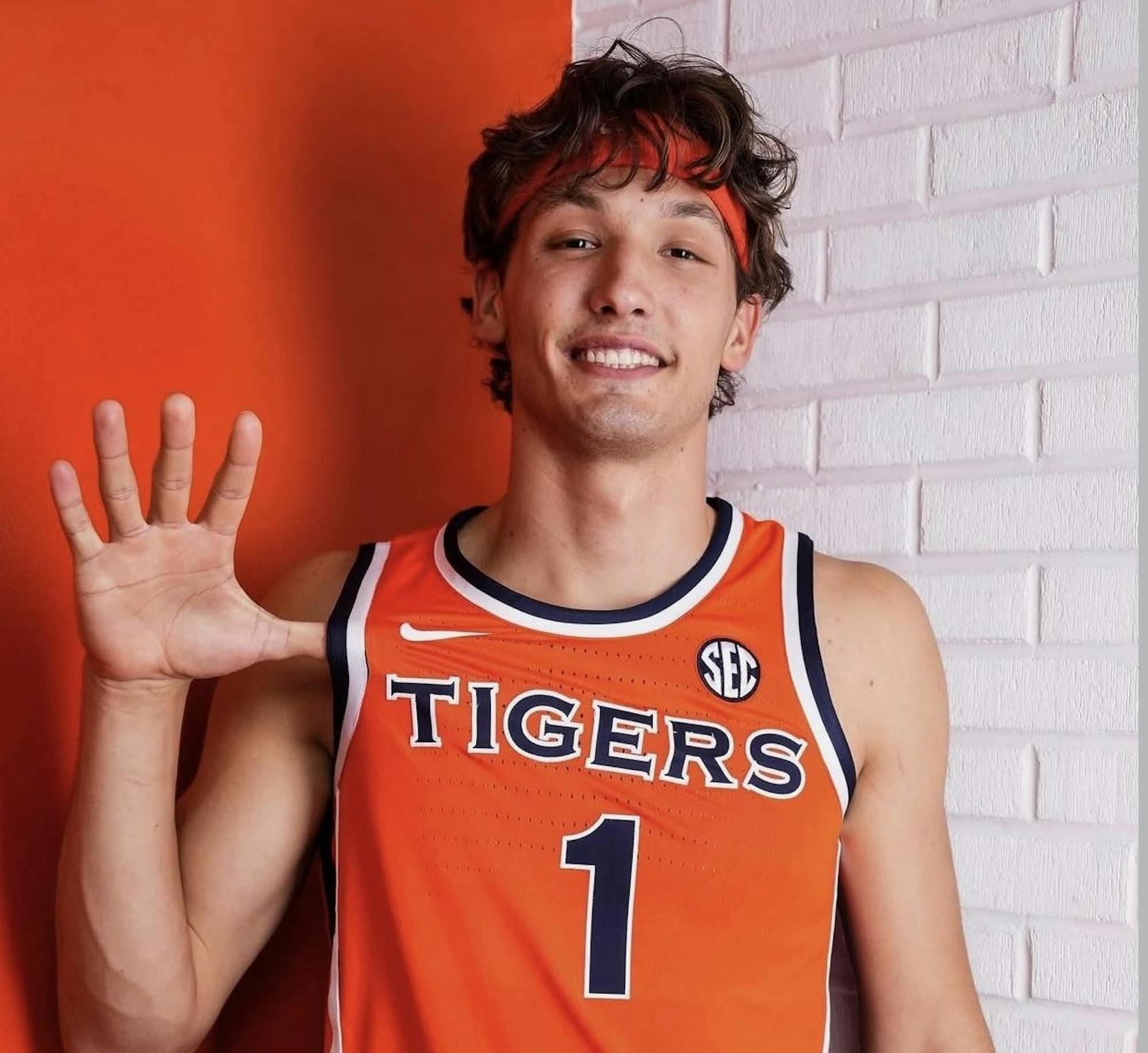 A young man in an orange basketball jersey with the word "TIGERS" smiles and holds up one hand with five fingers extended.