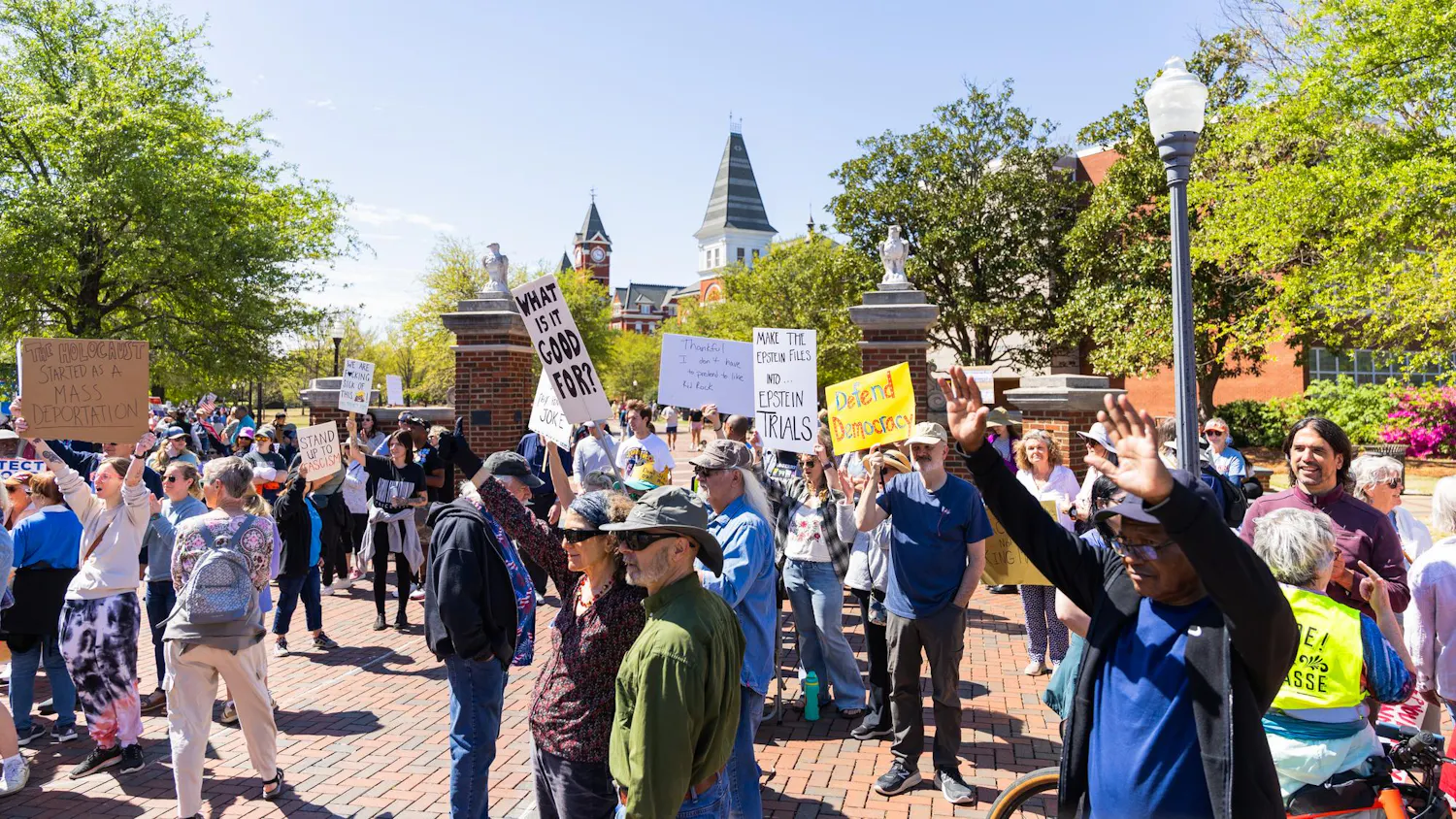 A crowd gathers outdoors, holding signs that address social issues, with trees and a historic building in the background.