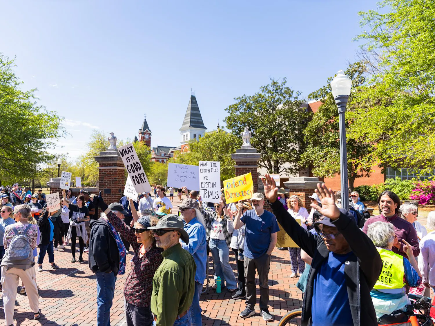 A crowd gathers outdoors, holding signs that address social issues, with trees and a historic building in the background.