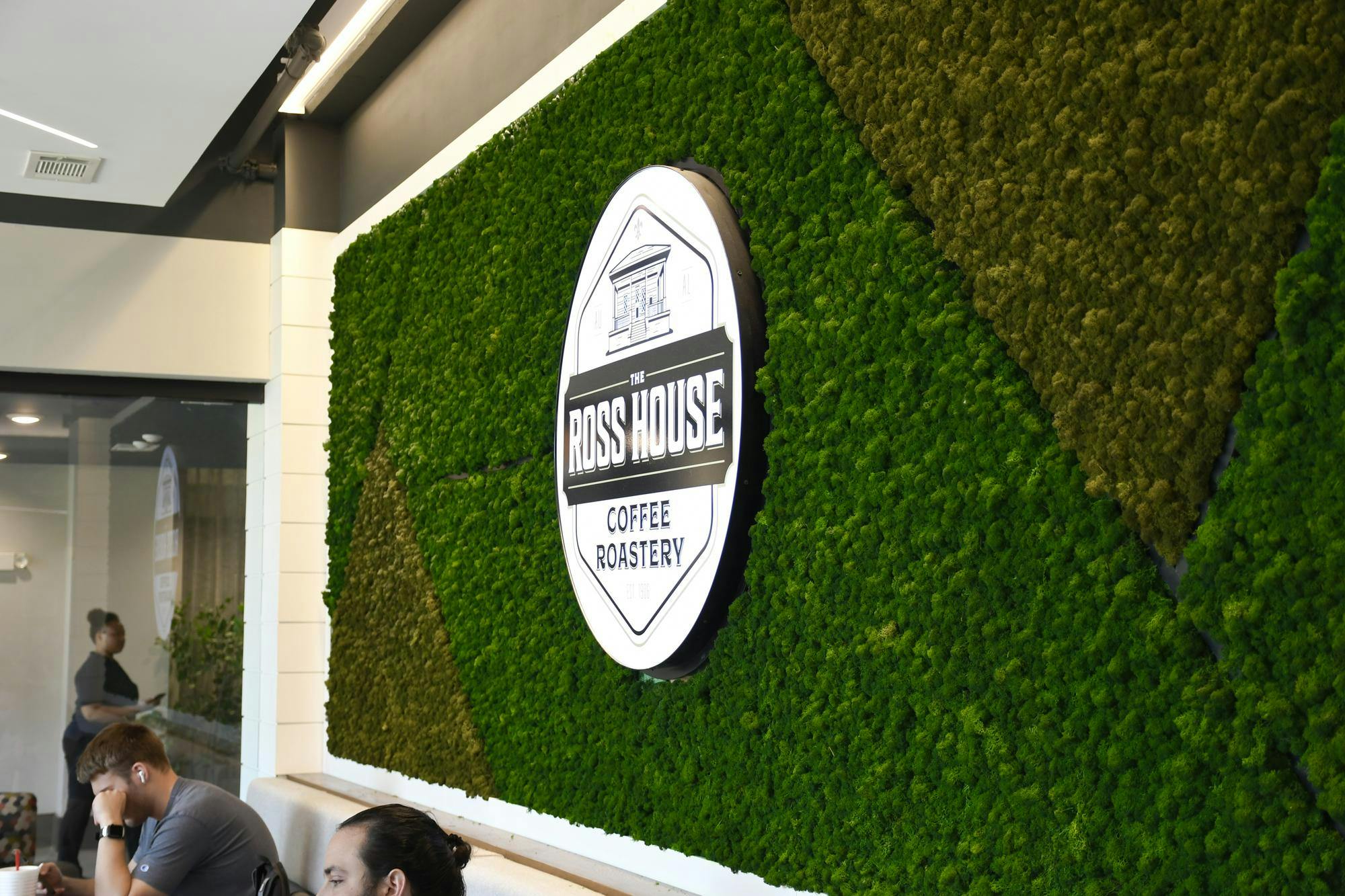A green moss wall features a circular sign reading "The Ross House Coffee Roastery," with people seated nearby.
