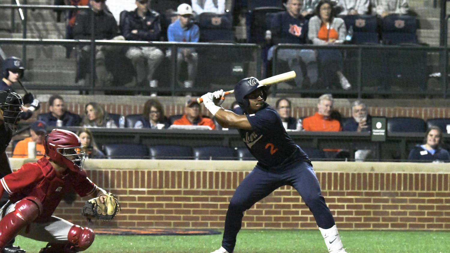 A baseball player stands ready to bat, while a catcher crouches in front, with spectators visible in the background.