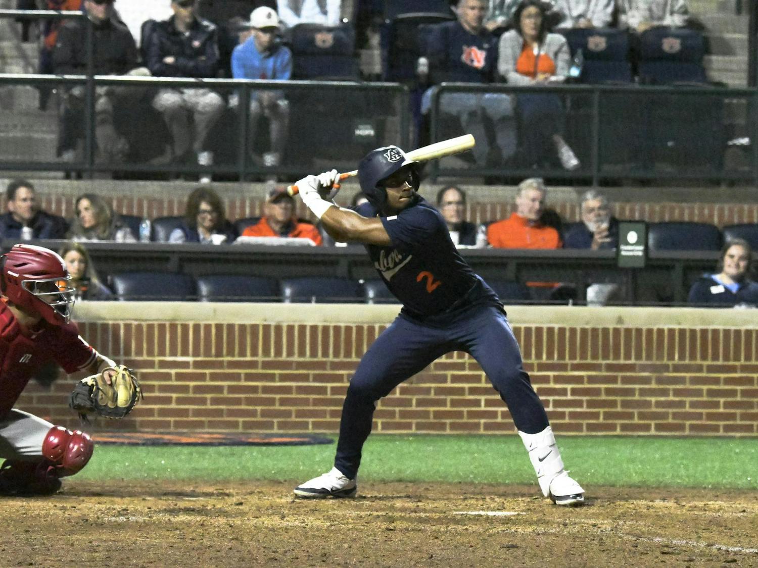A baseball player stands ready to bat, while a catcher crouches in front, with spectators visible in the background.