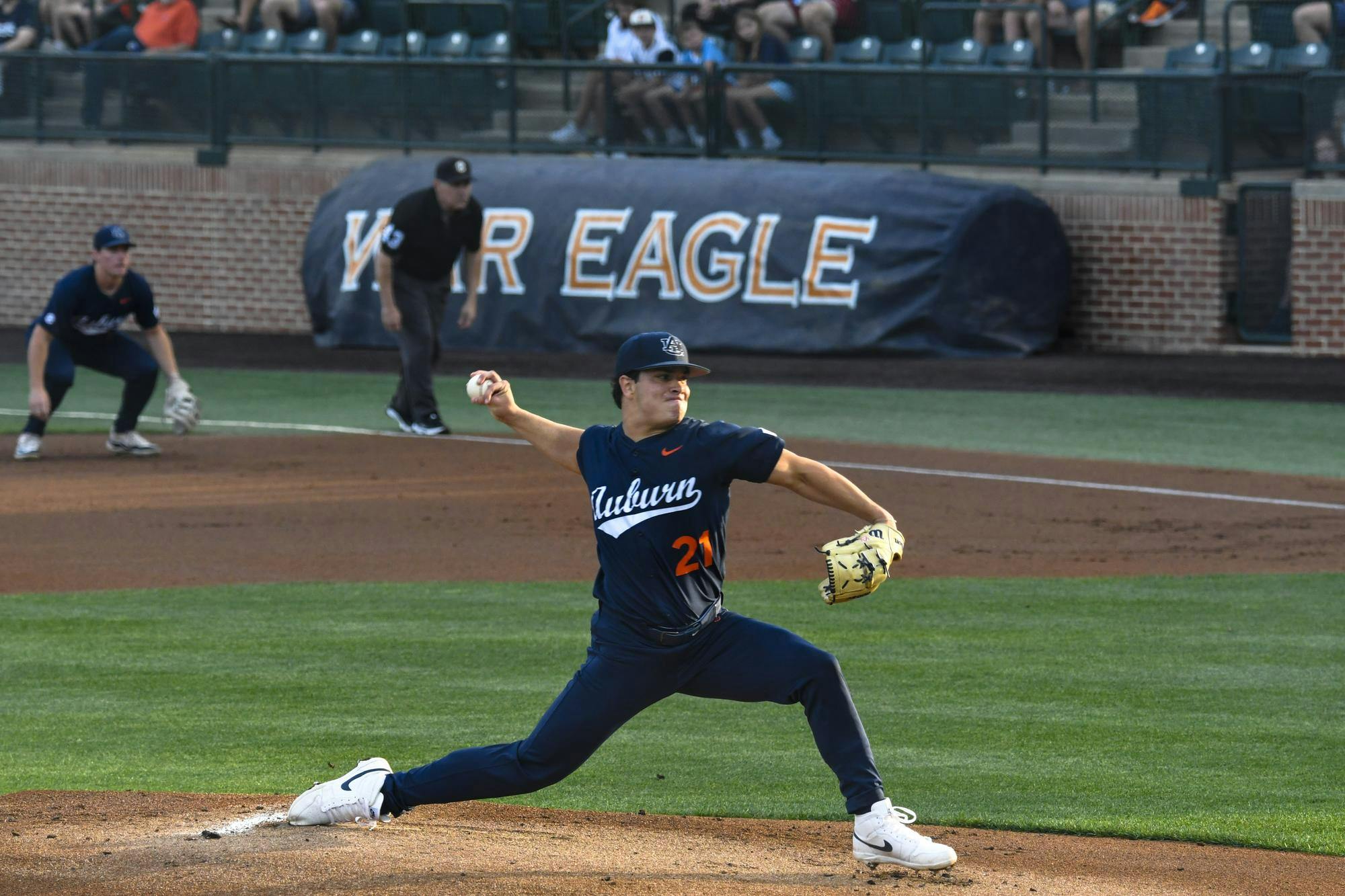 A baseball pitcher in a dark blue uniform winds up to throw a ball, with players and spectators in the background.