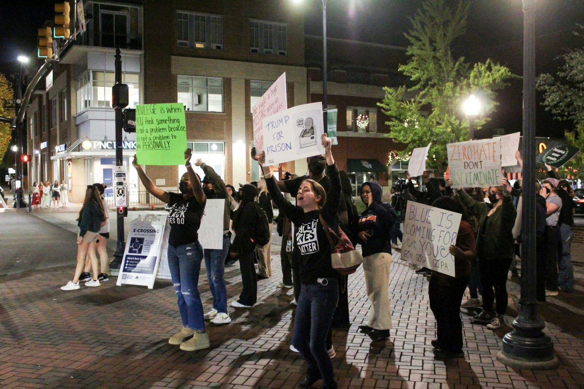 A group of people is gathered at night, holding signs with various political messages, while some are chanting.
