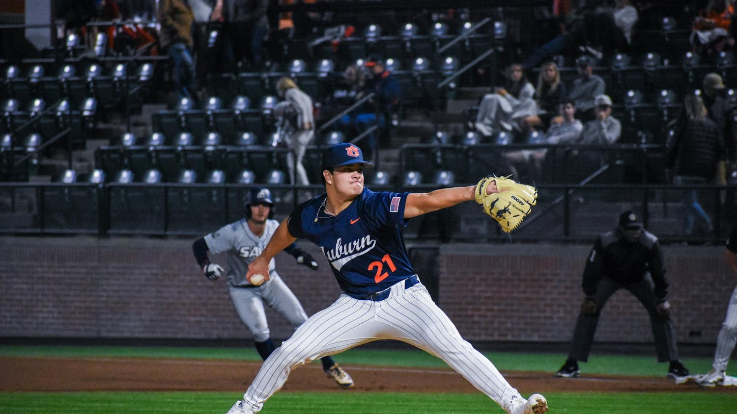 A baseball player in a blue jersey is in a pitching stance on the mound, while a runner in a gray uniform heads towards first base.