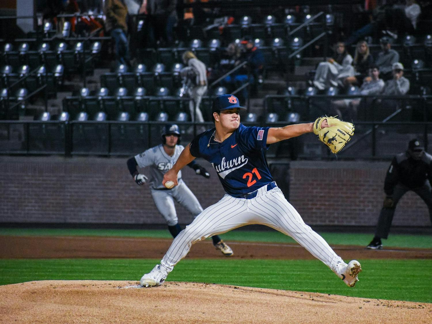A baseball player in a blue jersey is in a pitching stance on the mound, while a runner in a gray uniform heads towards first base.