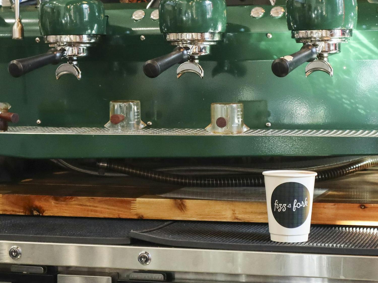 A large green espresso machine is positioned above a wooden surface with a paper cup labeled "fizz & fork" nearby.