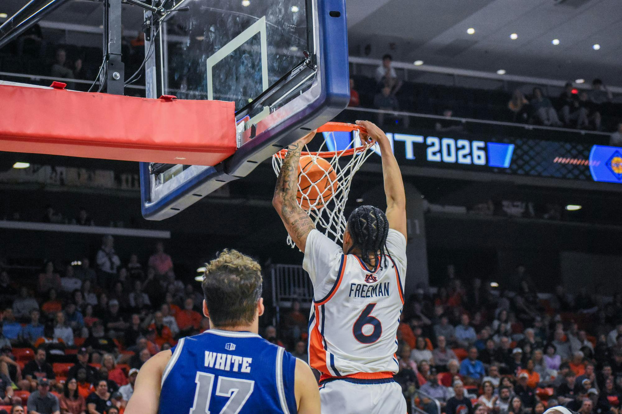 A player in a white jersey dunks a basketball into the hoop, while another player in a blue jersey watches closely.