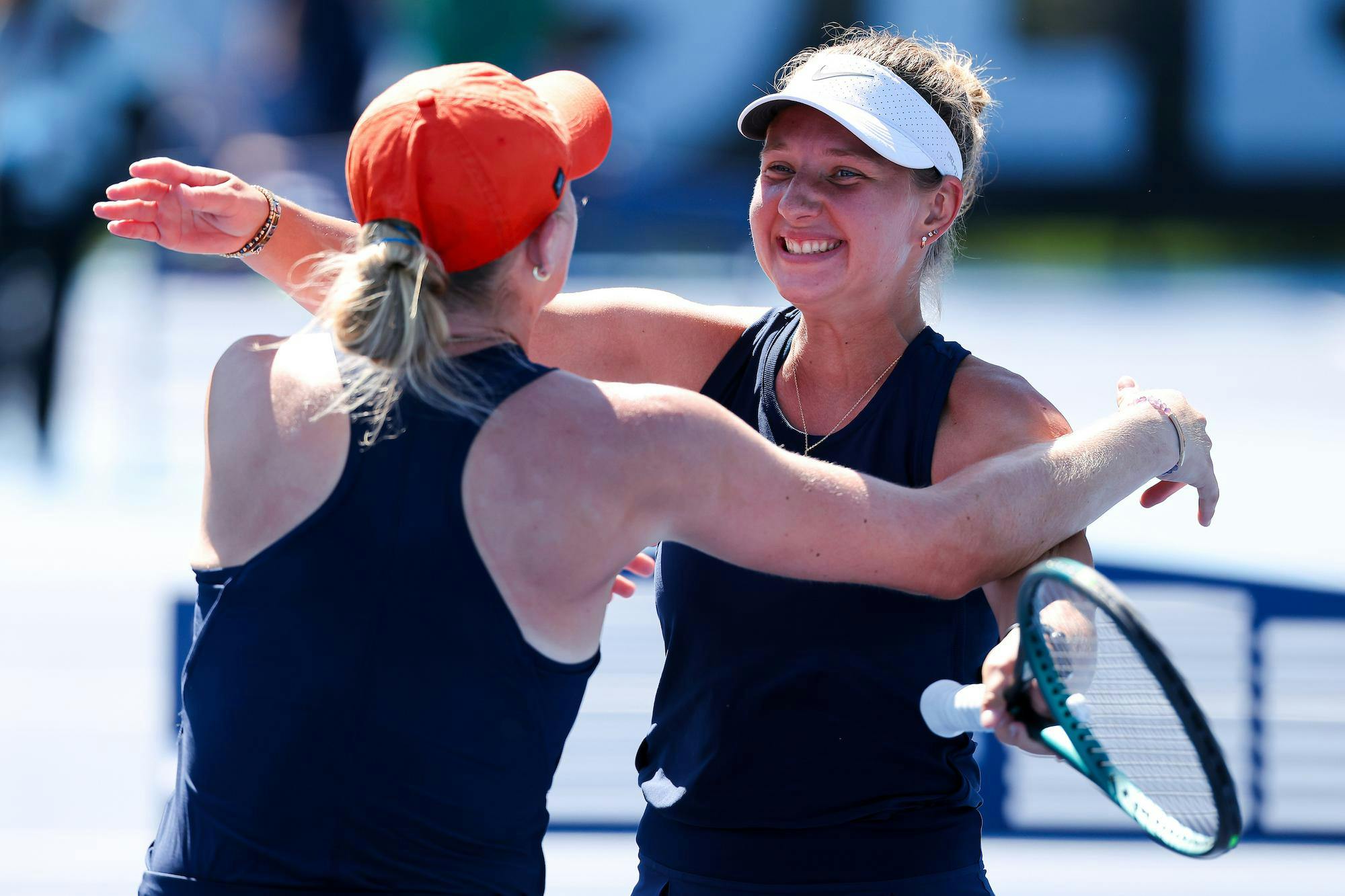 Two women, wearing matching sports outfits, embrace each other joyfully while one holds a tennis racket.