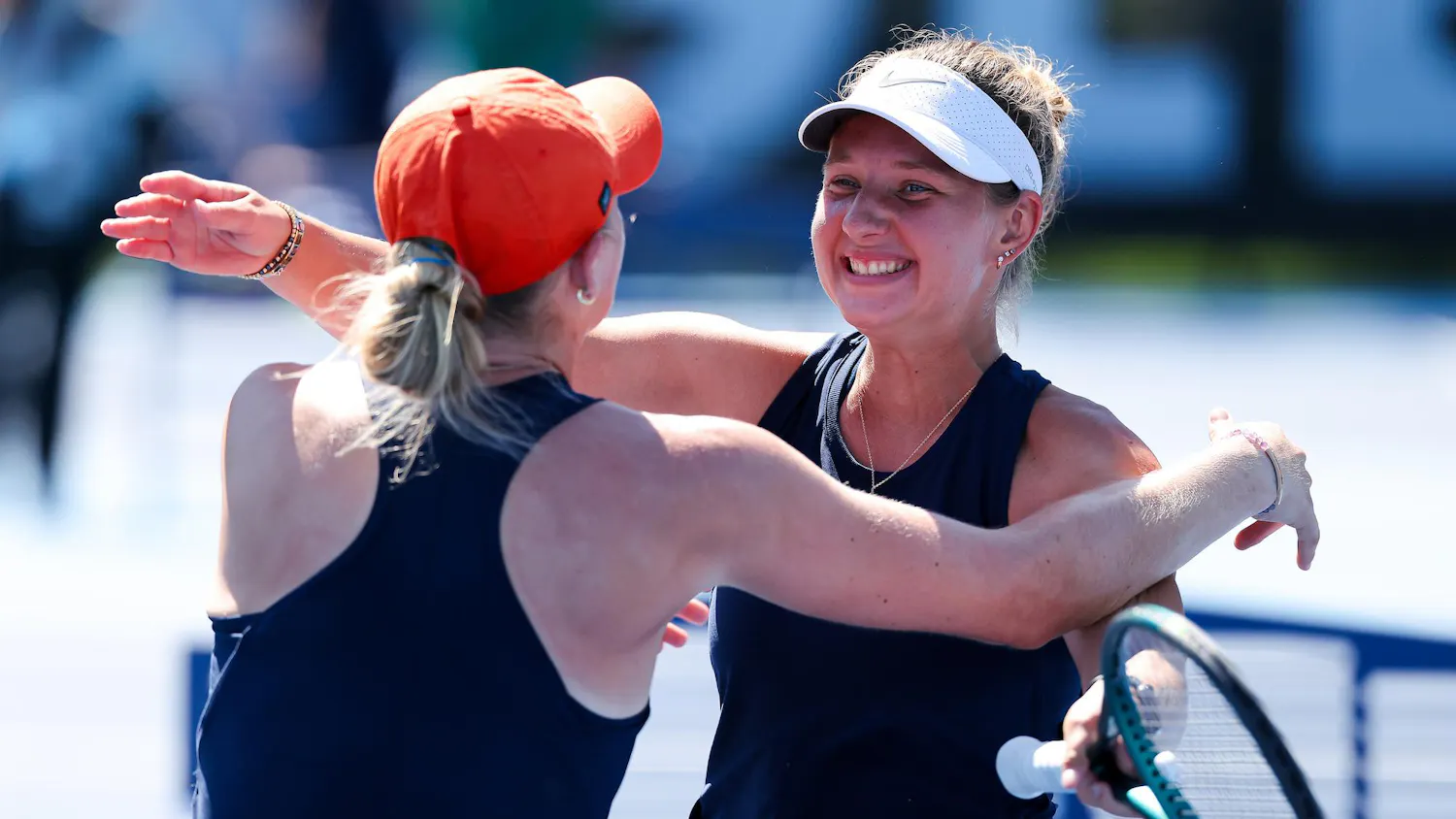 Two women, wearing matching sports outfits, embrace each other joyfully while one holds a tennis racket.
