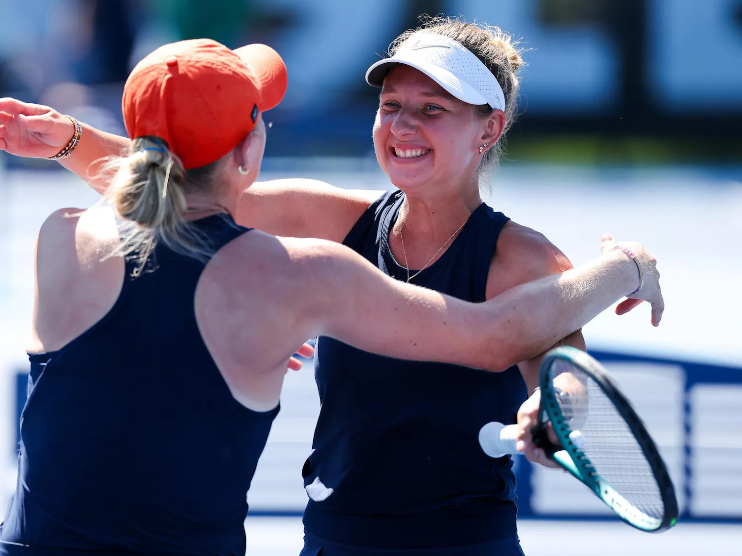 Two women, wearing matching sports outfits, embrace each other joyfully while one holds a tennis racket.