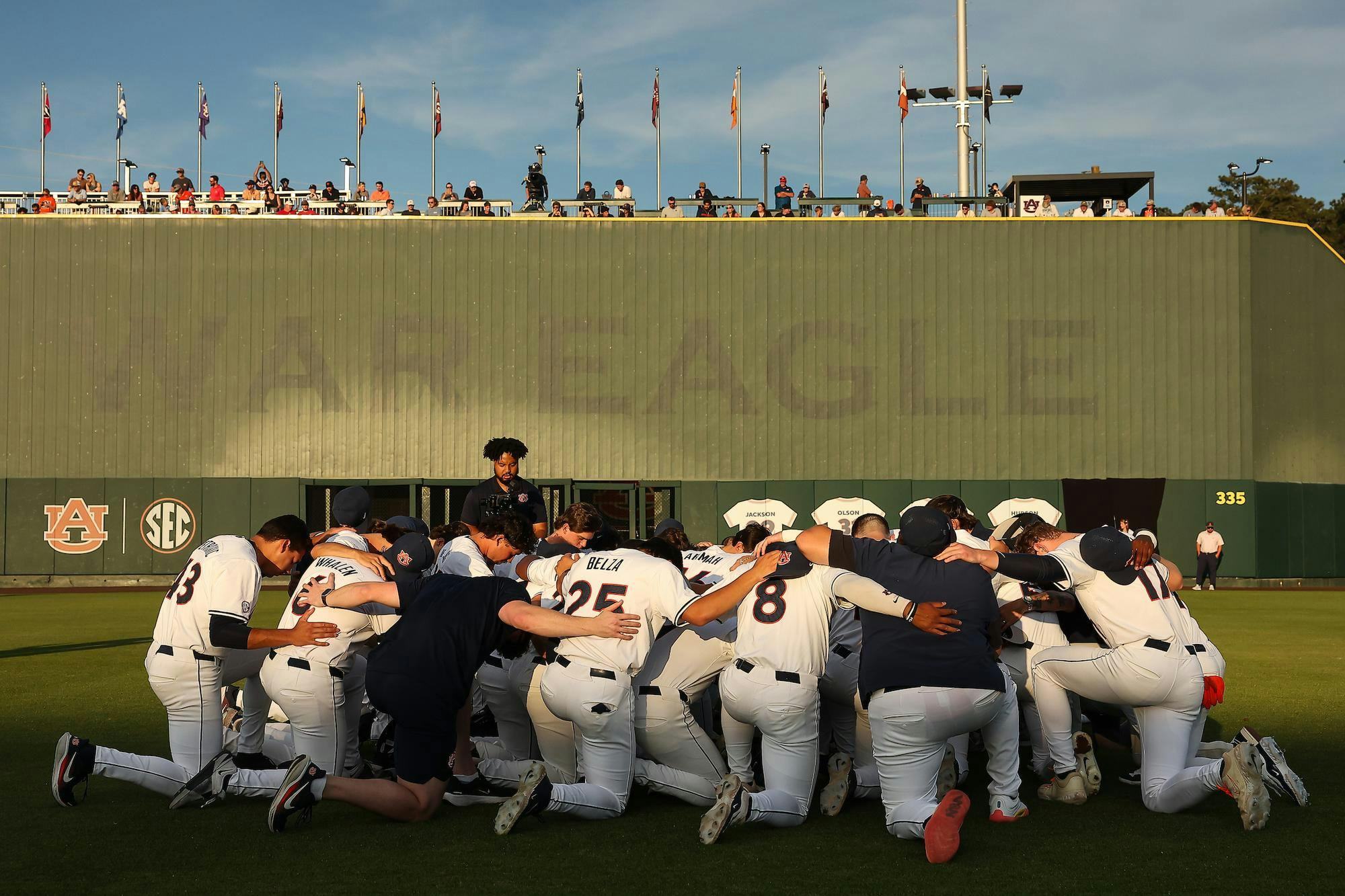 A baseball team kneels together in a huddle with a green scoreboard and spectators in the background.