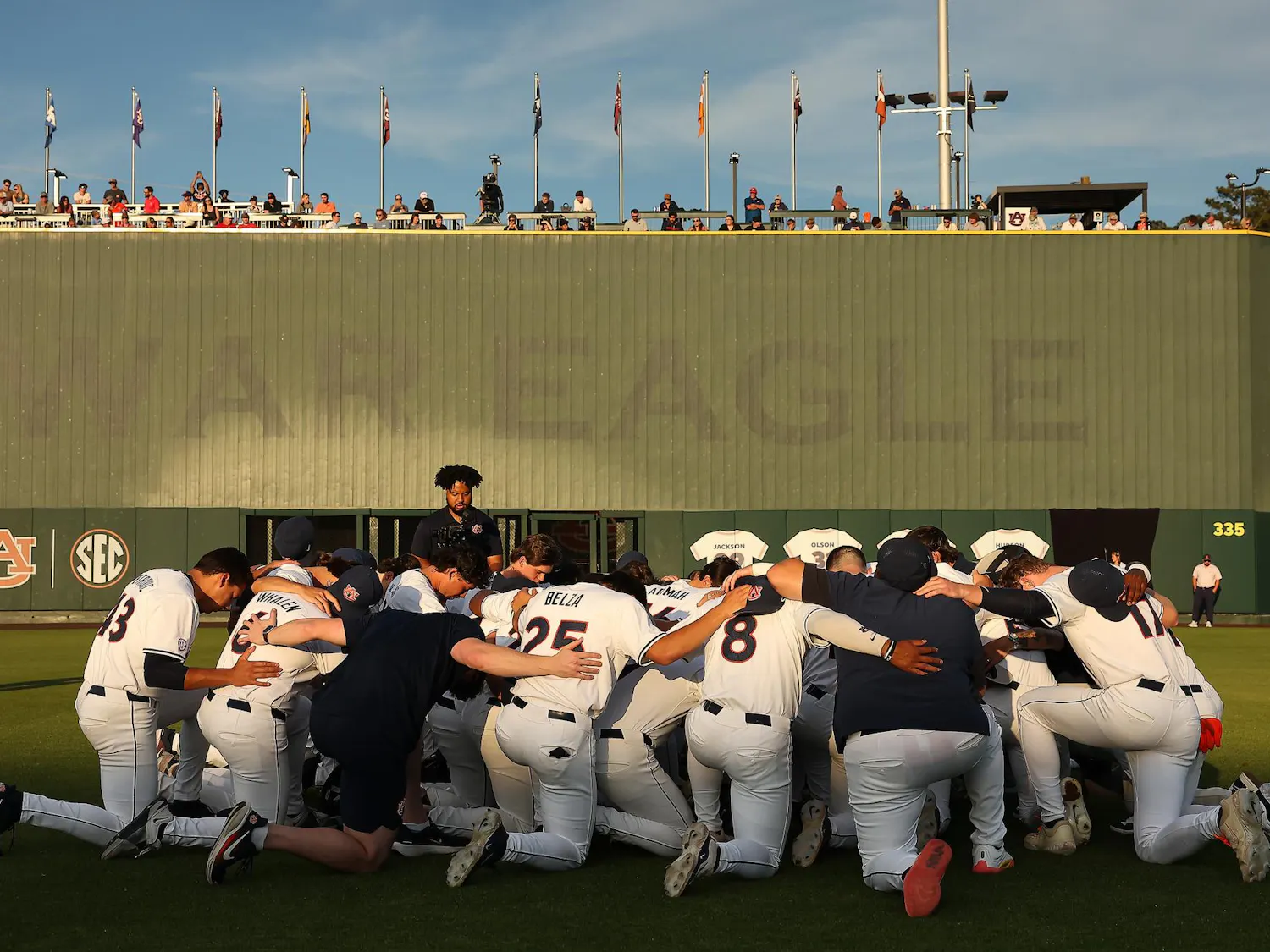A baseball team kneels together in a huddle with a green scoreboard and spectators in the background.