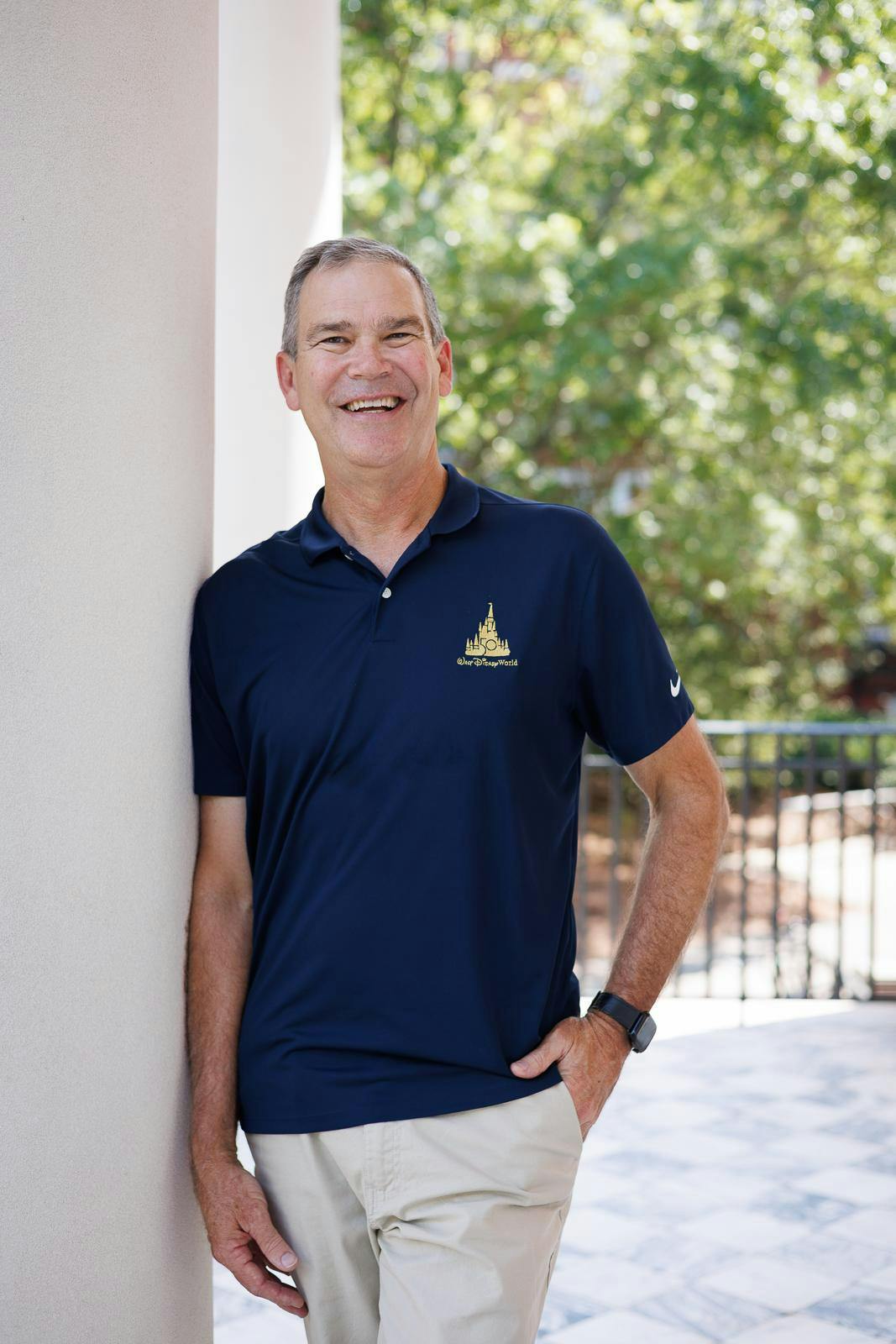 A smiling man in a navy polo shirt stands casually against a column outdoors, surrounded by trees and natural light.