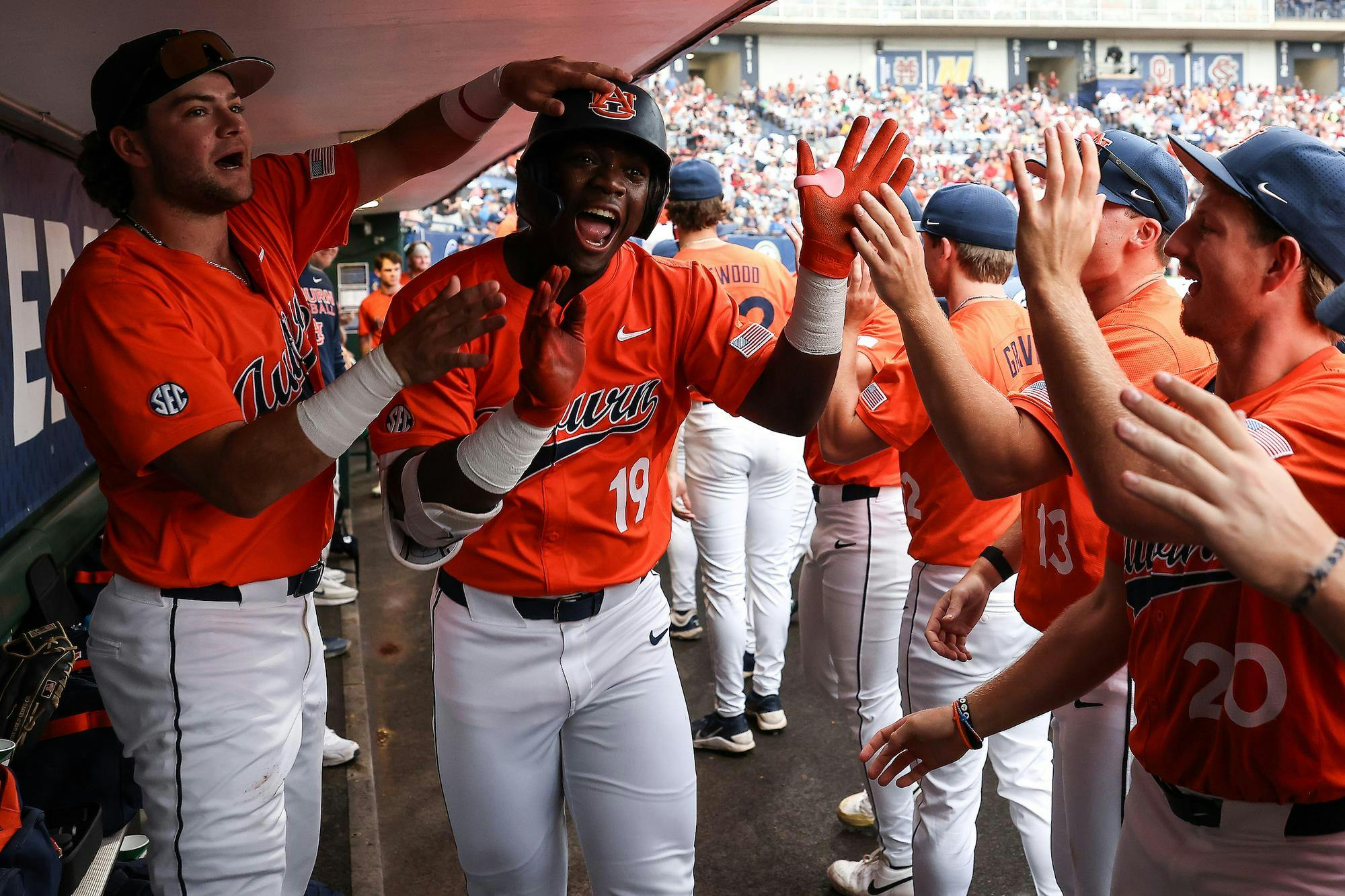Players in orange baseball jerseys celebrate in a dugout, clapping and cheering as one player smiles broadly.