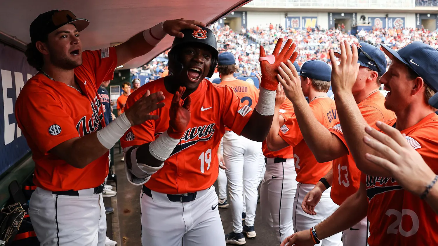 Players in orange baseball jerseys celebrate in a dugout, clapping and cheering as one player smiles broadly.