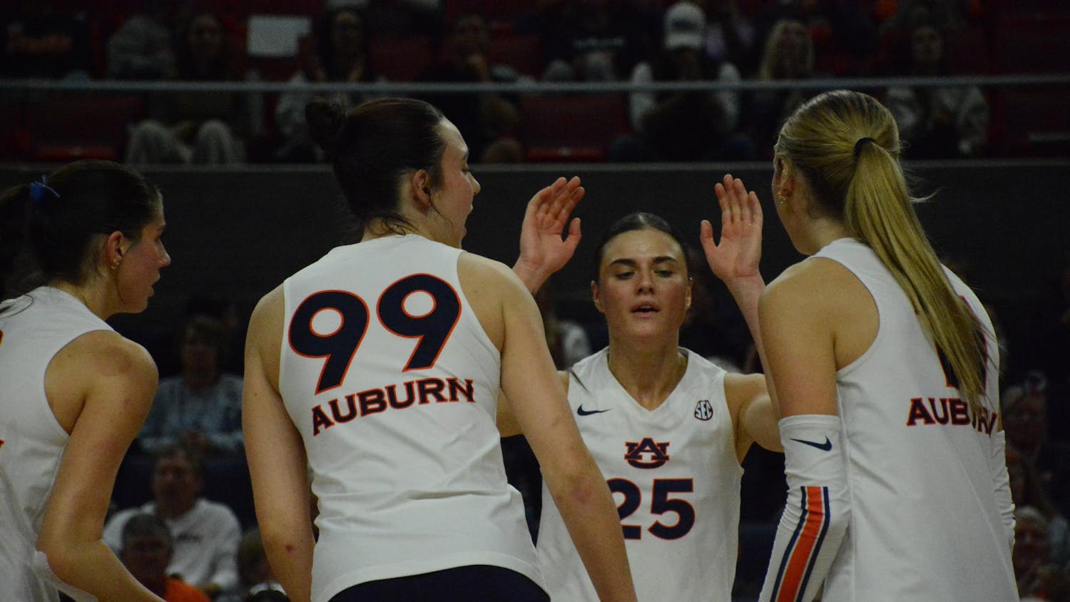 Four female volleyball players in white uniforms are engaged in a celebratory moment, with one raising her hands while others have their backs turned.